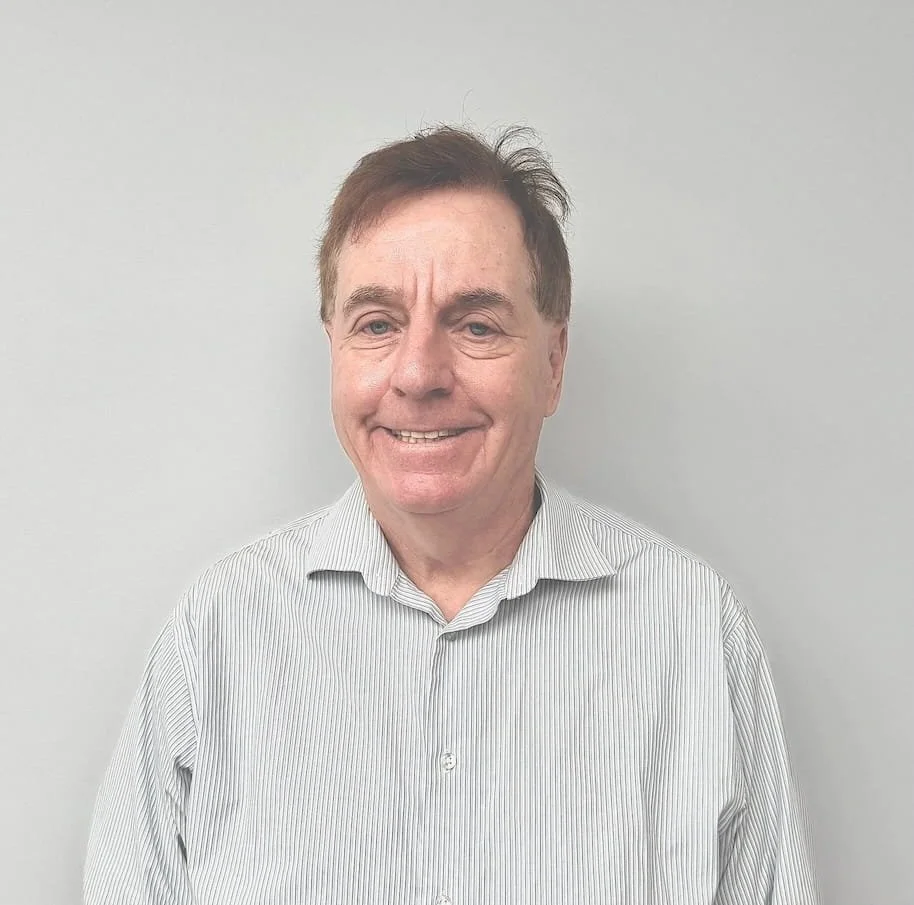 Photograph of a person wearing a striped button-up shirt standing against a plain light-colored wall. The person's face is obscured.