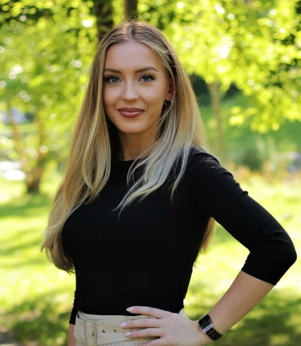 A person with long blonde hair wearing a white coat with a name tag that reads 'Weronika, Neurofeedback Technician' standing outdoors in a park with green trees in the background.