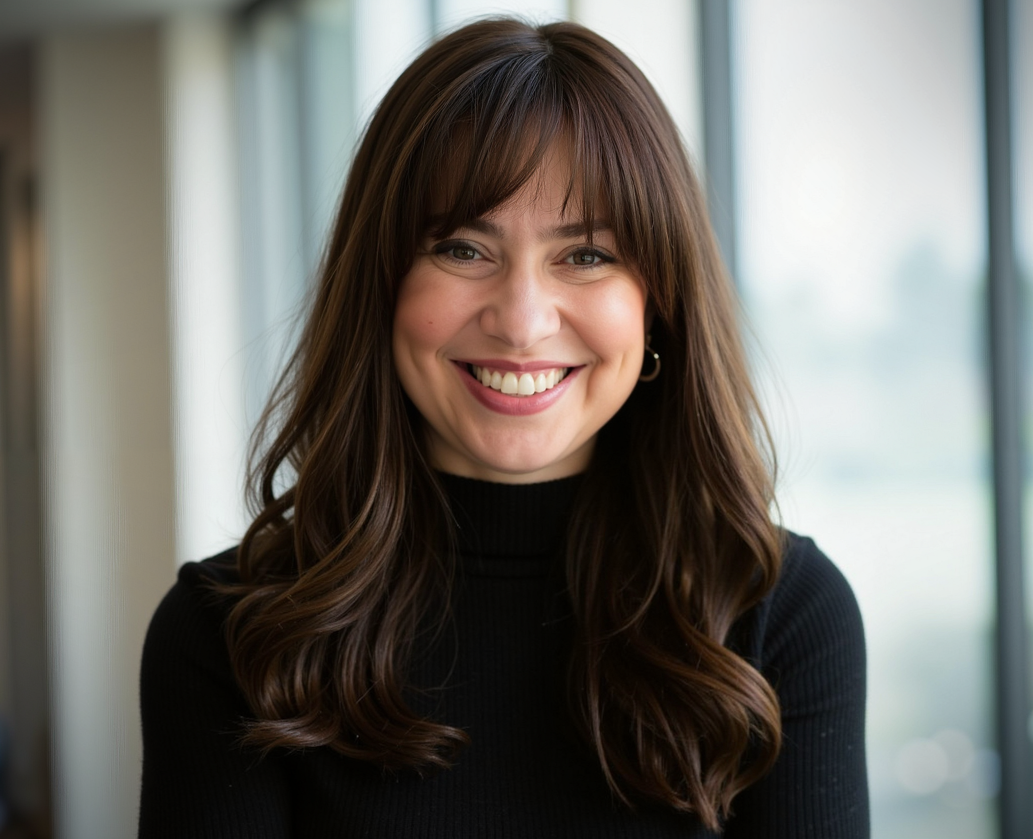 A woman with long, wavy brown hair and bangs, wearing a black turtleneck, smiling at the camera in front of a window with a blurred outdoor background.