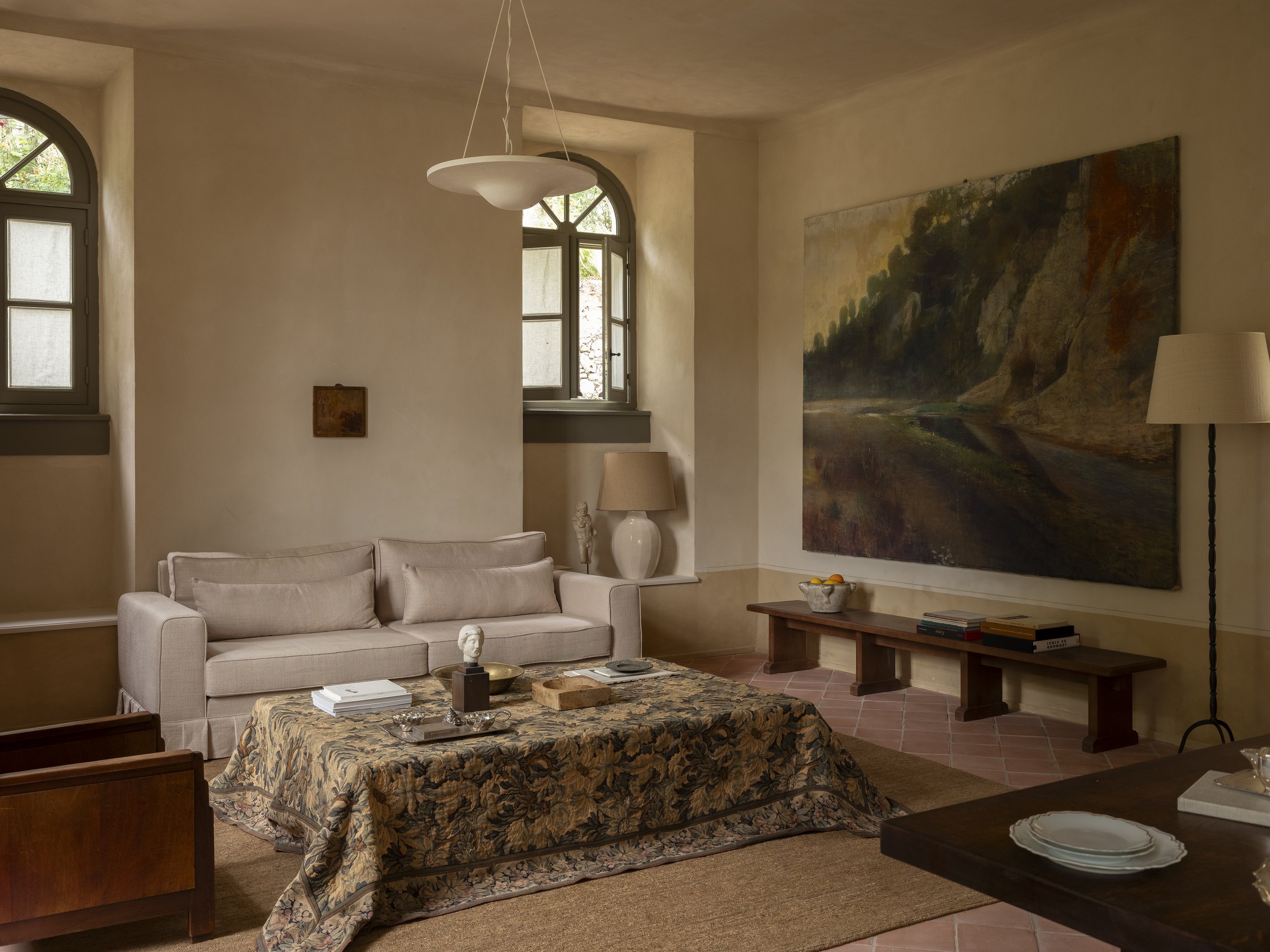 Living room with a beige sofa, wooden coffee table covered with a tapestry, large landscape painting on the wall, and two windows with arched frames.