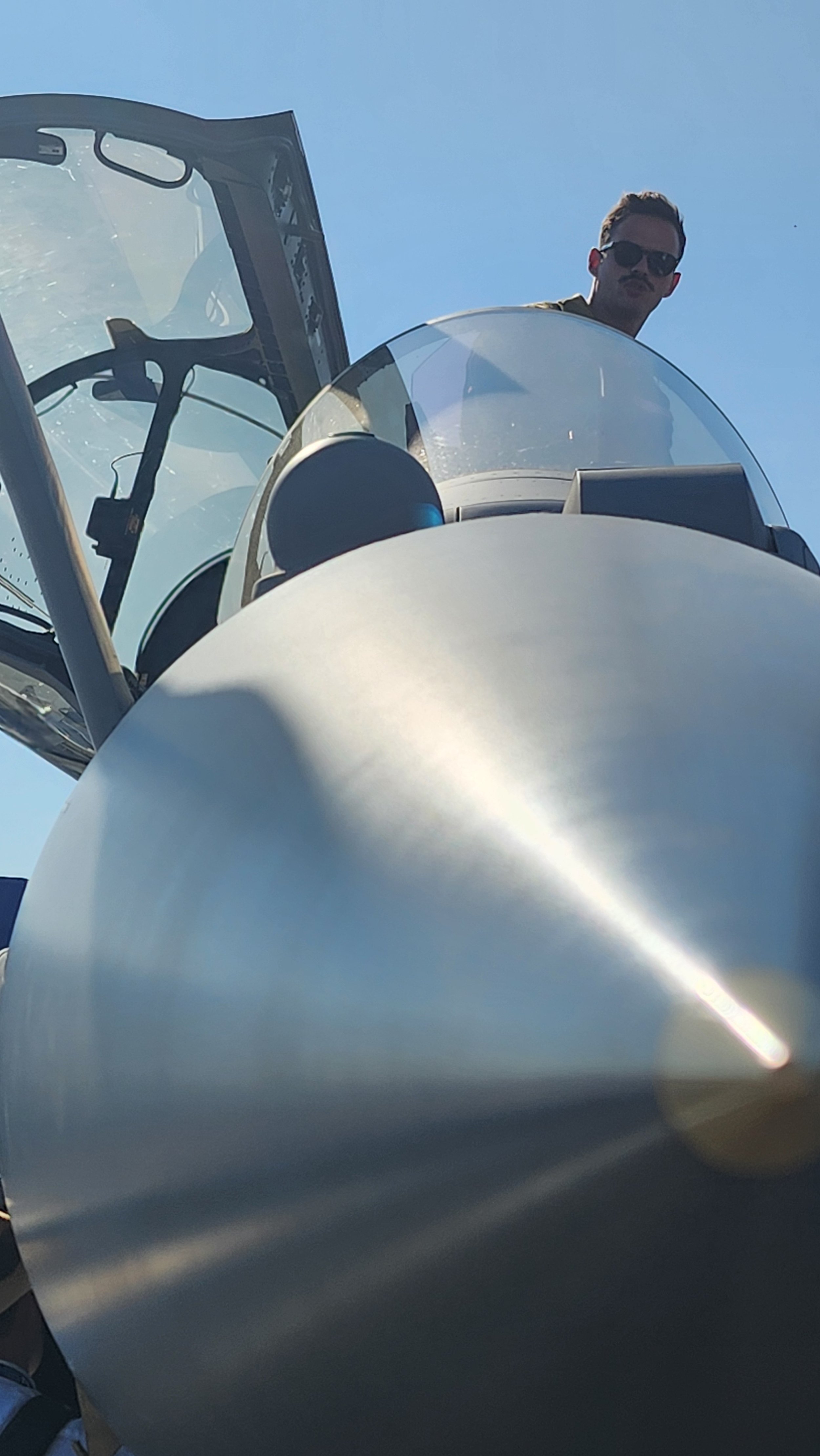 Un homme portant des lunettes de soleil est visible à l'intérieur d'un avion de chasse. L'avion est vu de dessous, avec un ciel bleu clair en arrière-plan.
