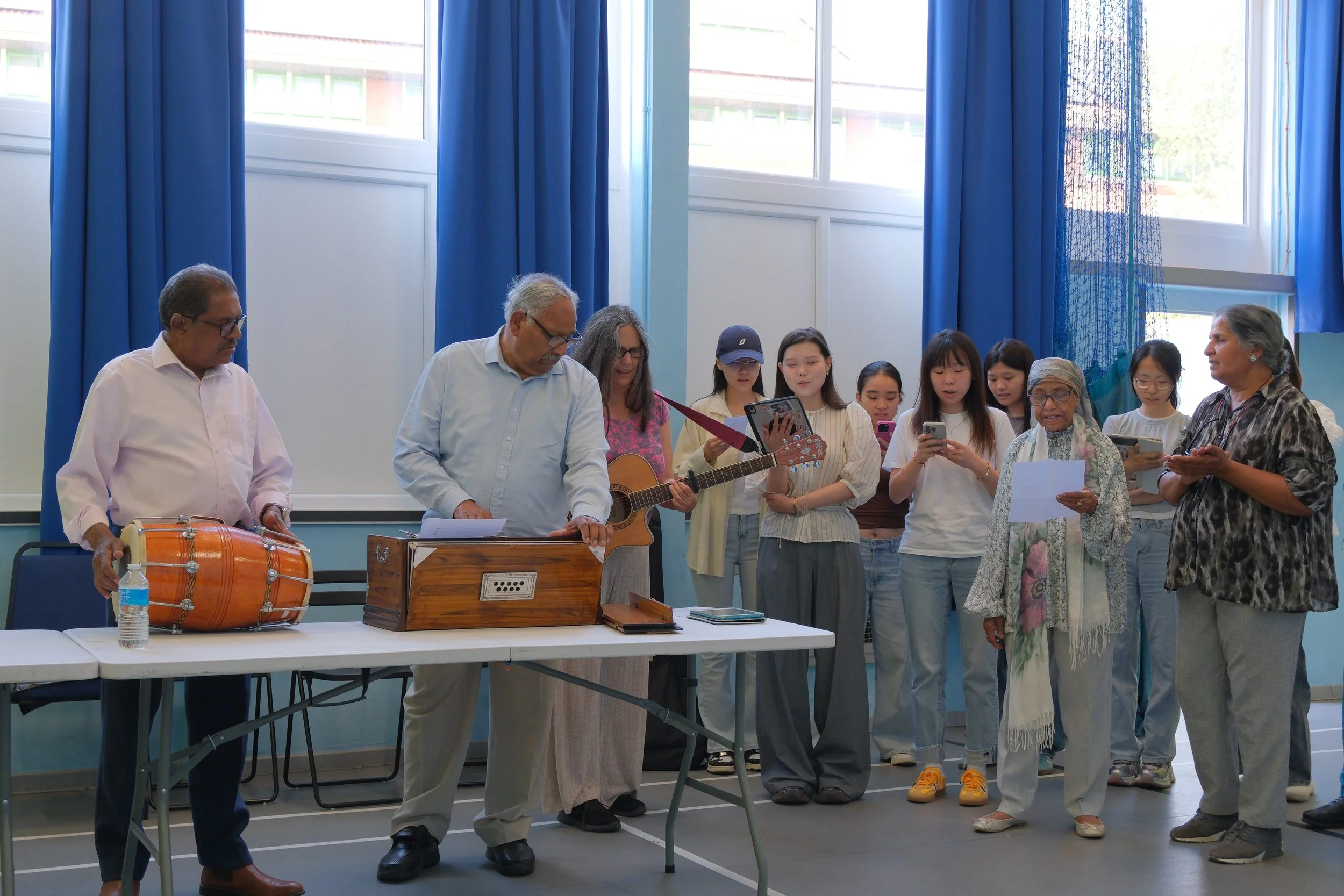 Group of people singing and playing musical instruments in a room with blue curtains.