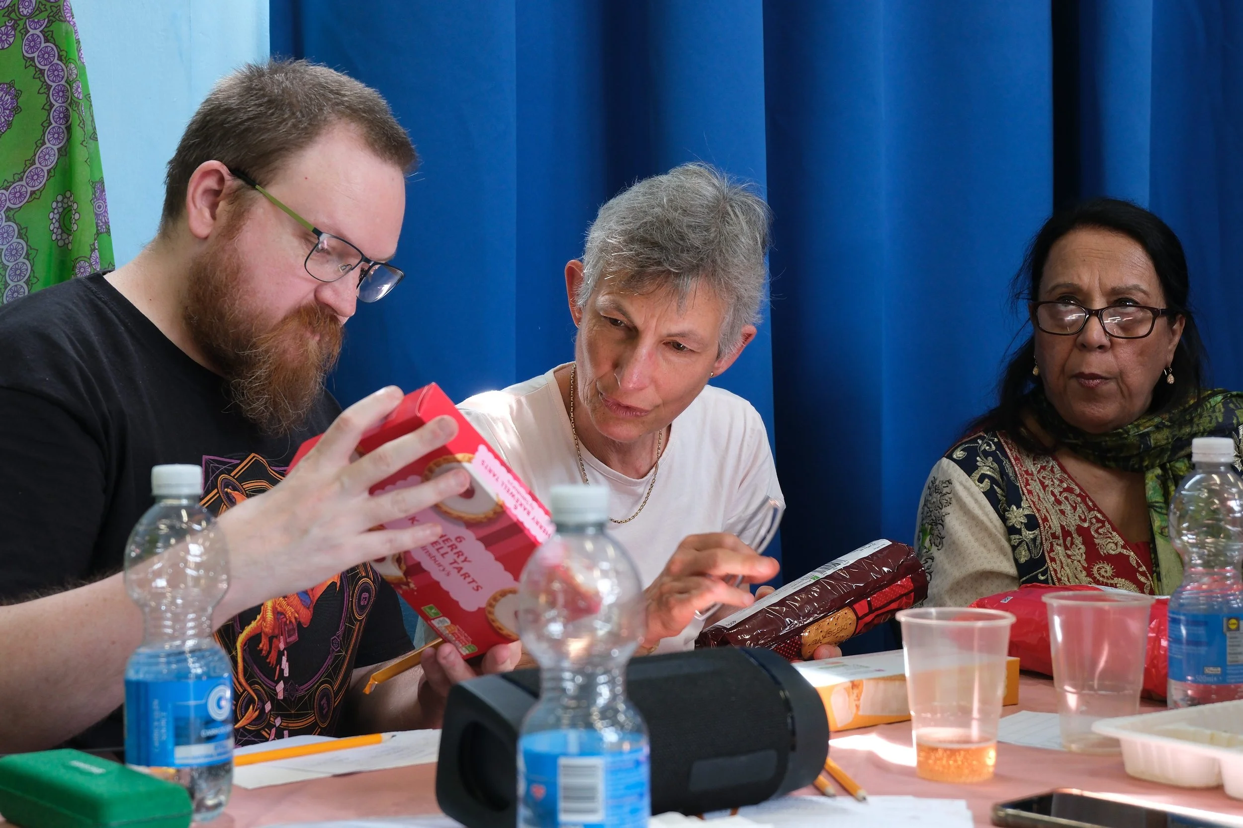 Three people sitting at a table with water bottles, snacks, and papers, engaged in a discussion or activity. Blue curtains in the background.