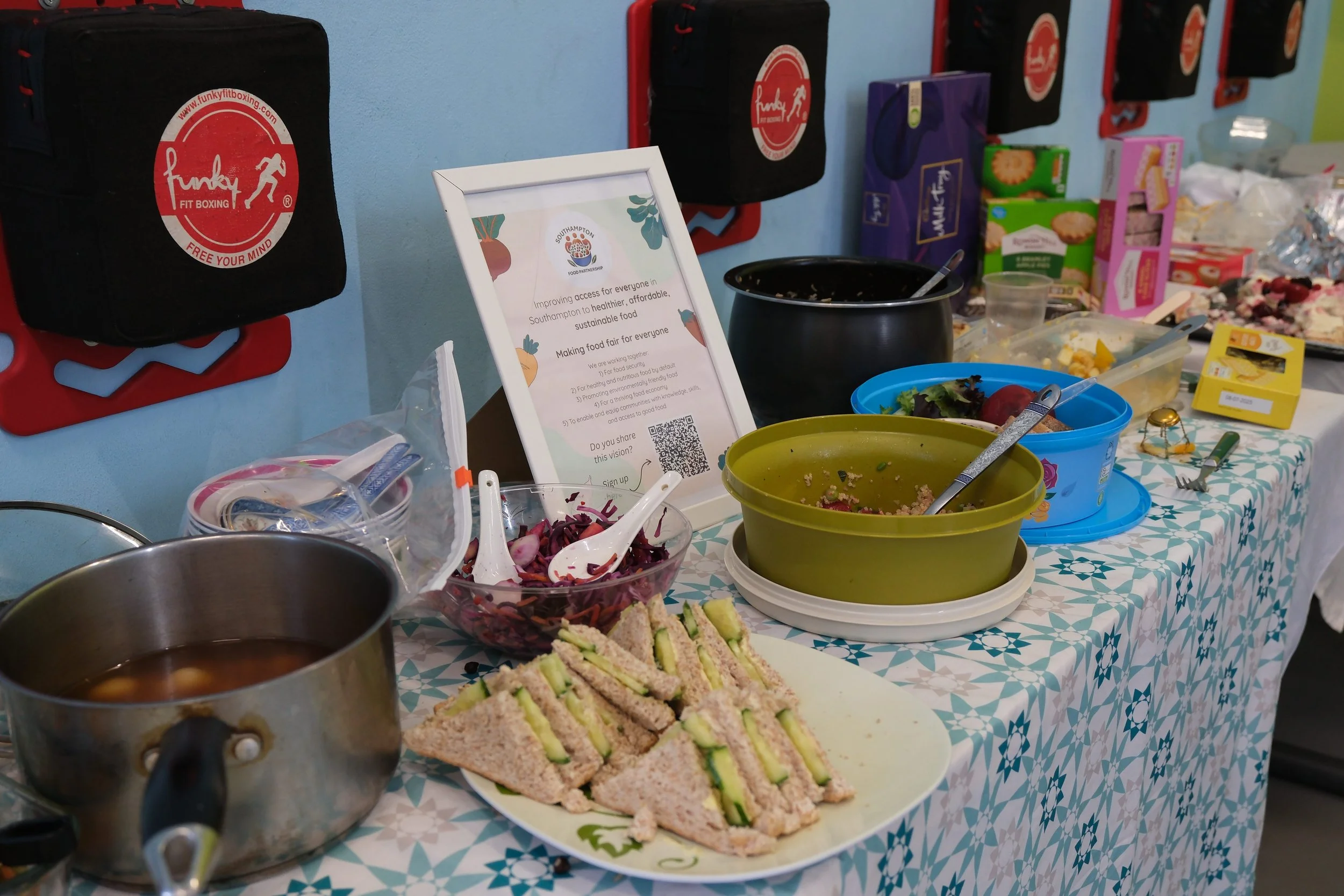 A table with various food items and containers, including salads, sandwiches, and dessert, set up for a community event or gathering.