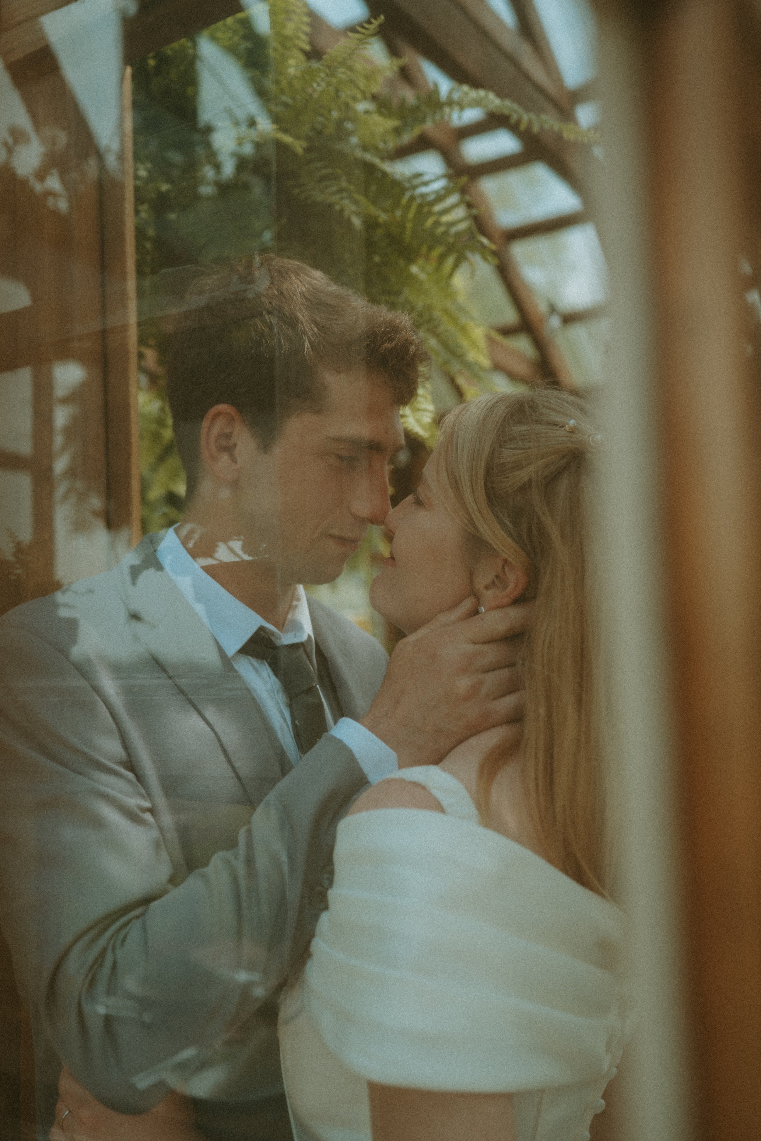 A man and woman embrace closely, about to kiss, through a glass window. The man is wearing a gray suit and black tie, and the woman has long blonde hair wearing a white outfit. There are green plants and wooden elements in the background.
