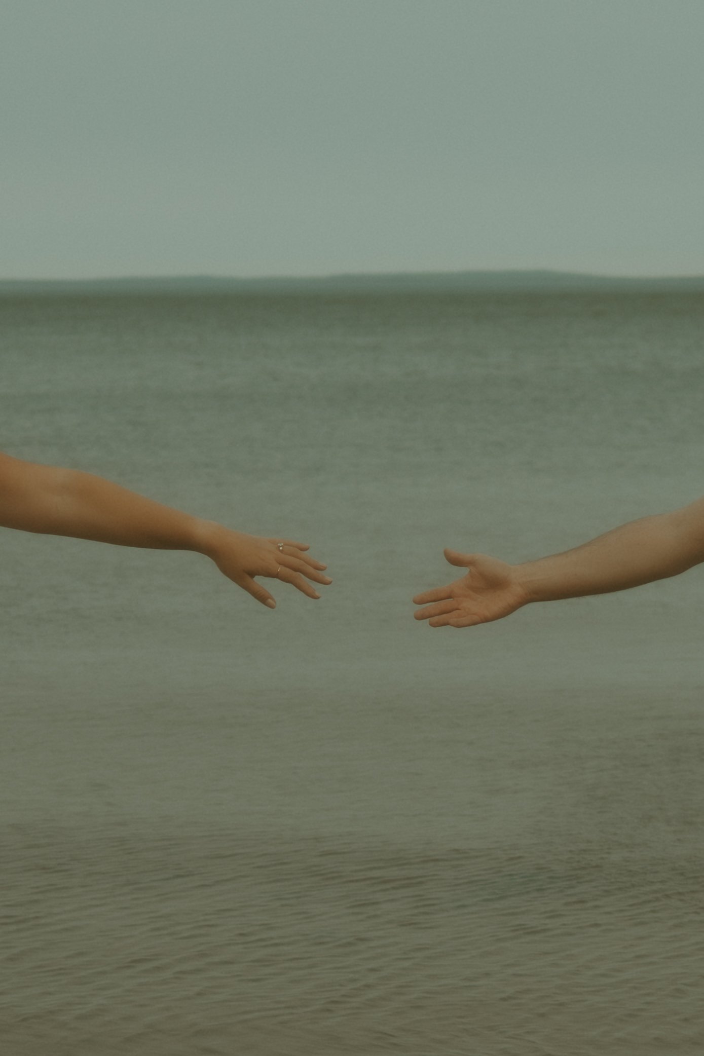 Two hands reaching out to each other over a body of water, with the horizon in the background.