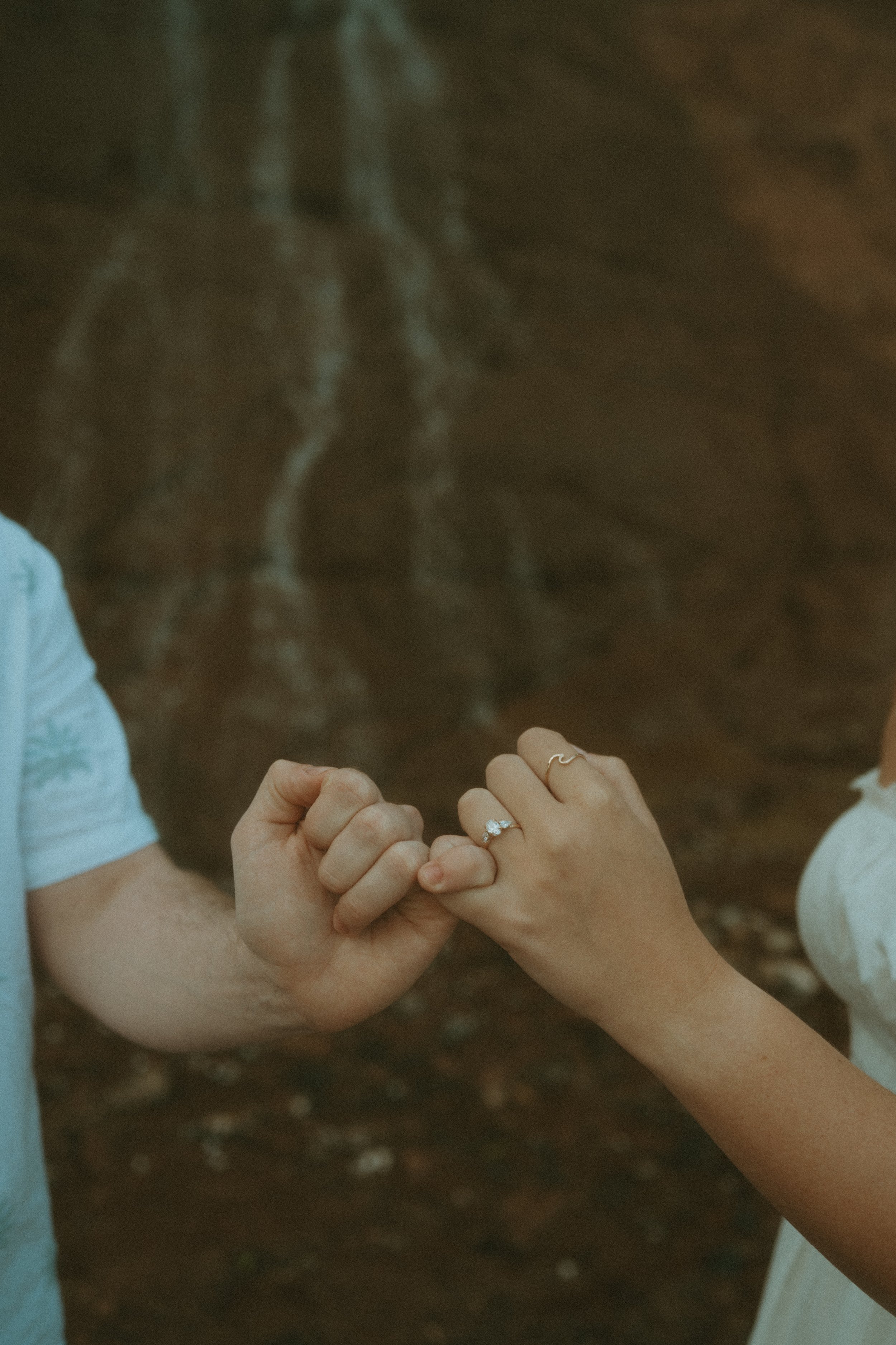 A close-up of two people holding hands, with the woman displaying an engagement ring with a large gemstone, holding hands in front of a rocky background.
