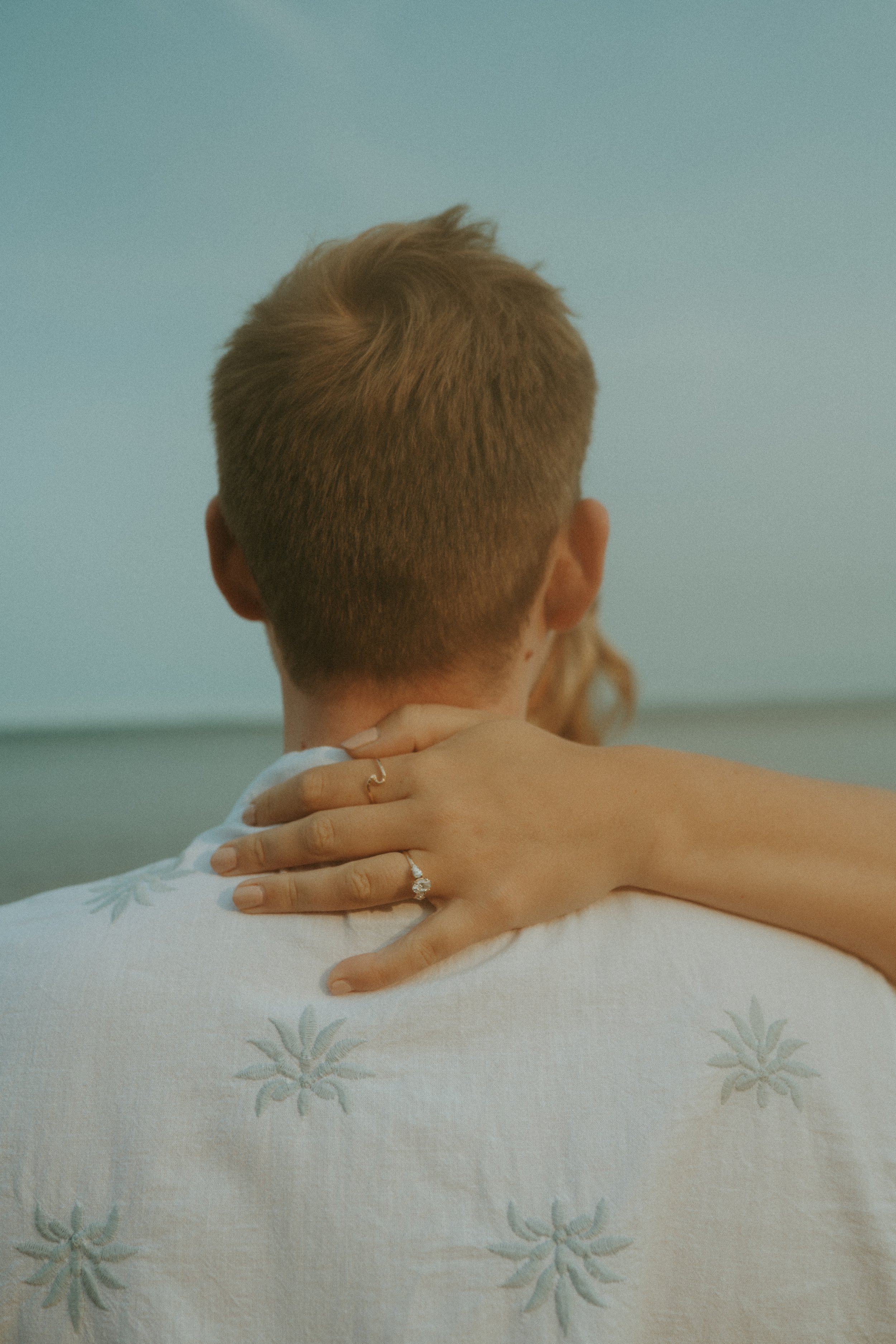 A person with short, light brown hair, seen from behind, with a womanβs hand resting on their shoulder, showcasing a wedding ring, in front of a light blue background.