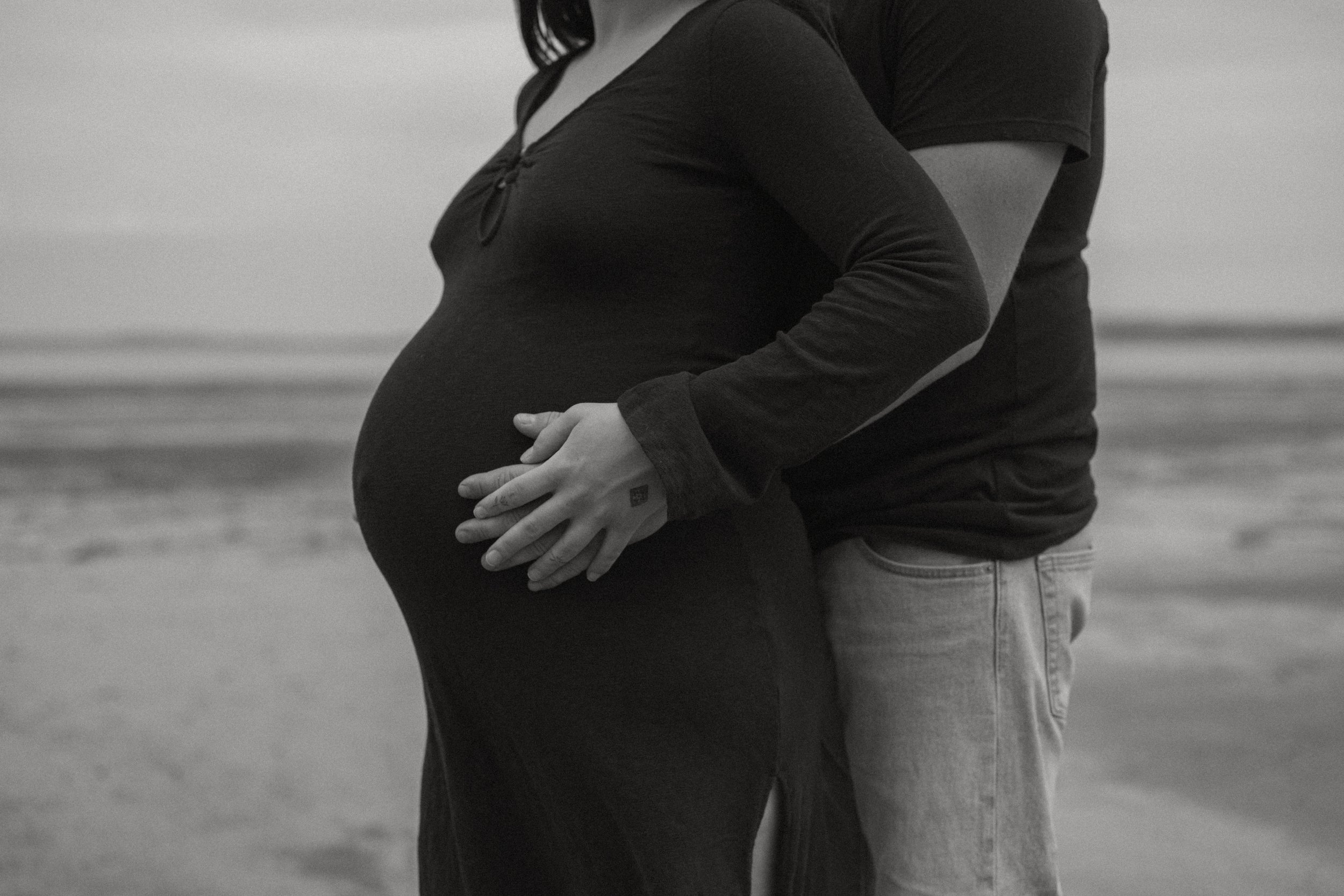 A black and white photo of a pregnant woman standing on a beach, with her partner embracing her from behind and holding her belly.