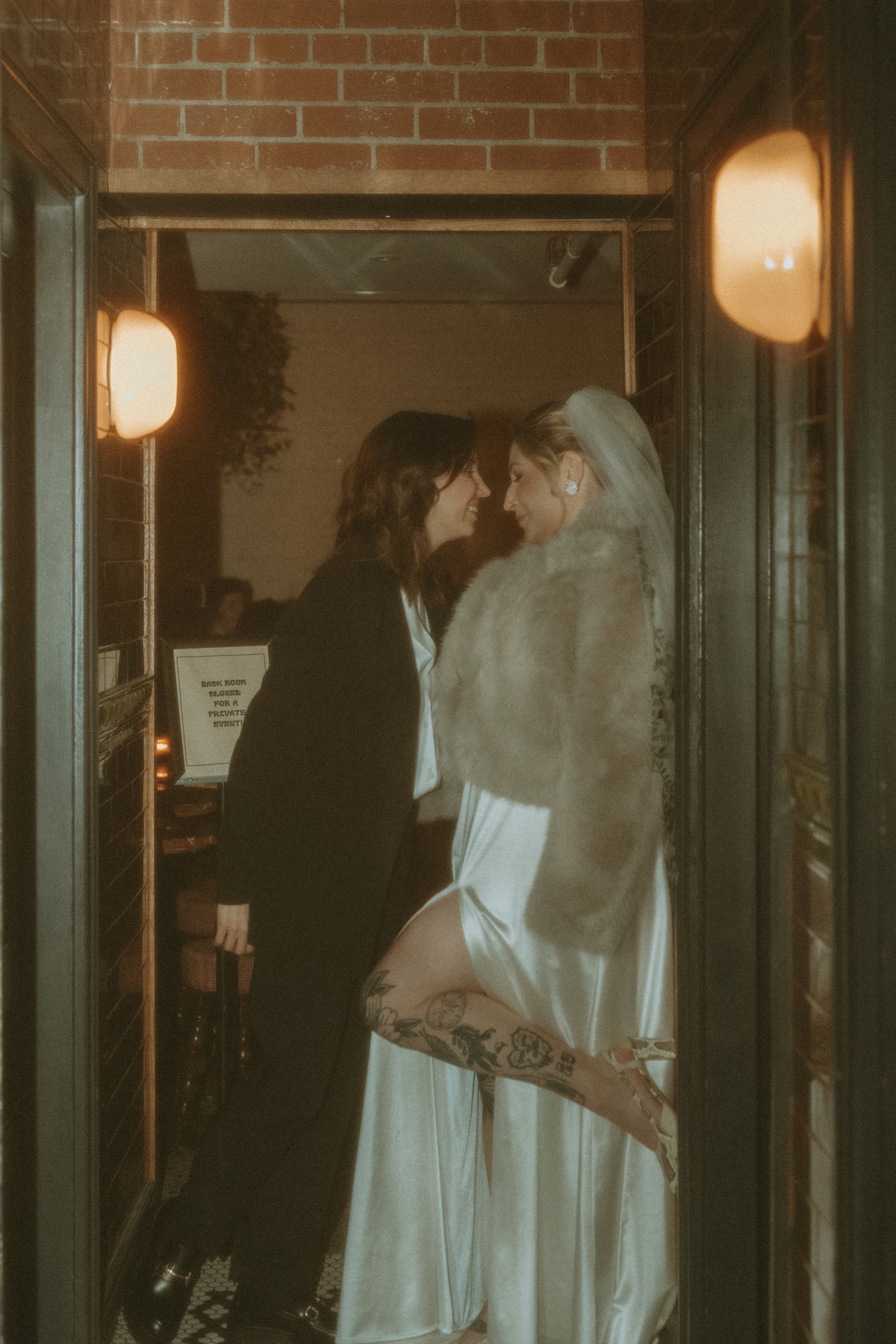 Two women, one in a tuxedo and one in a white satin wedding dress with a faux fur stole, are standing close together in a doorway. They are touching foreheads, smiling, and appearing happy. The woman in the wedding dress is lifting her leg, revealing
