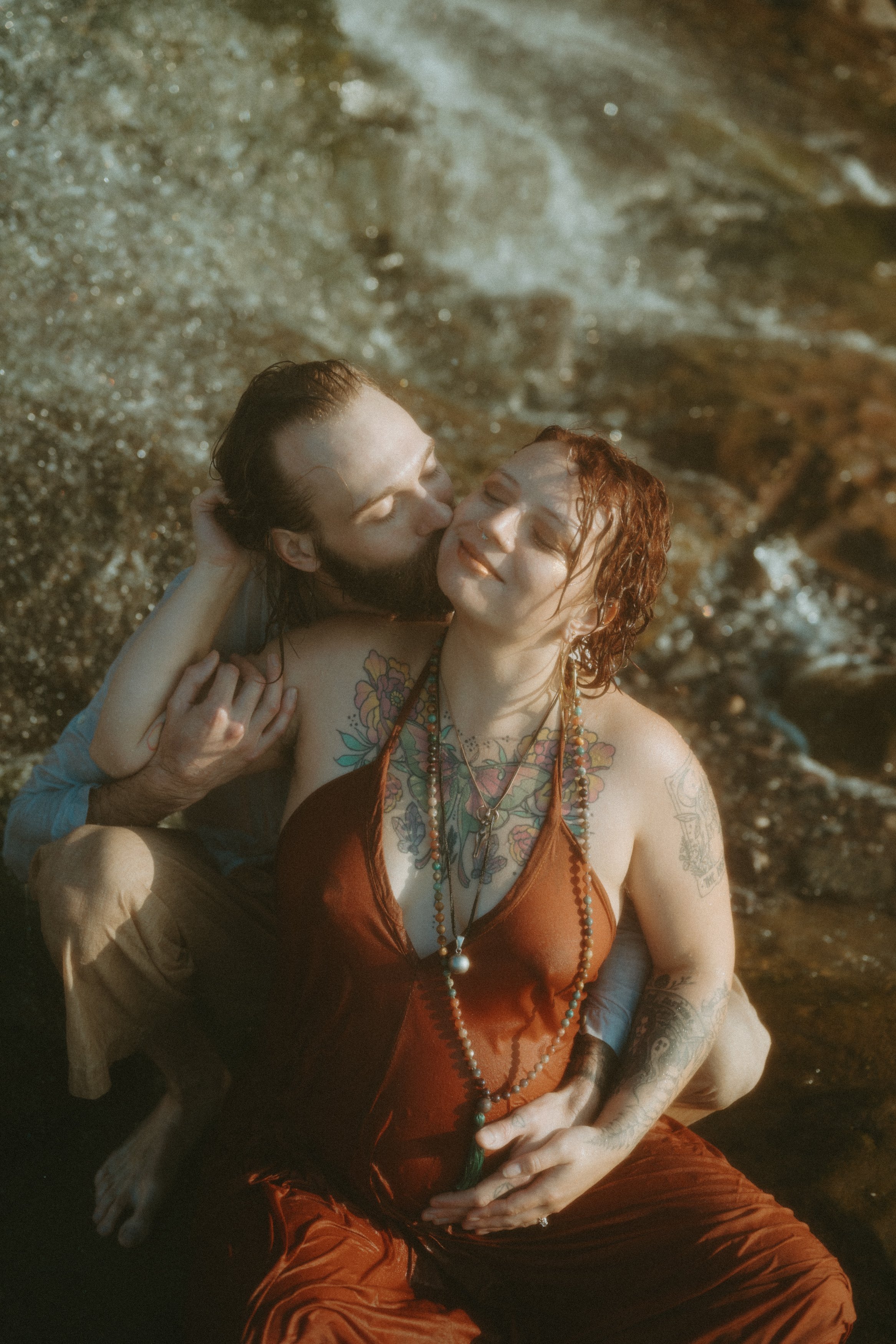 A man and woman joyfully sitting in a river, embracing and smiling, surrounded by rocks and water.