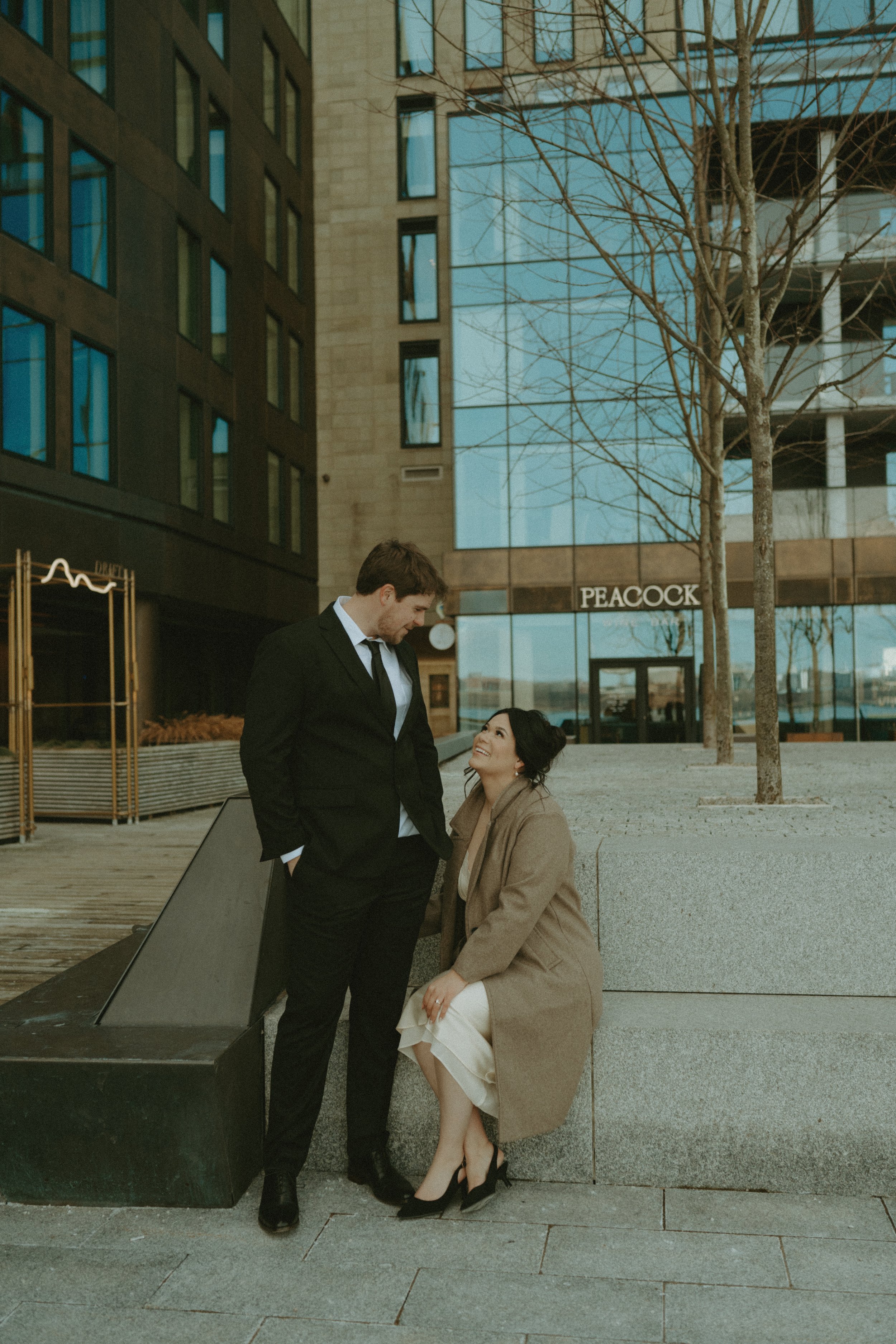 A man in a black suit and a woman in a beige coat and heels are smiling at each other outdoors in an urban area with glass buildings and a sign that says Peacock.
