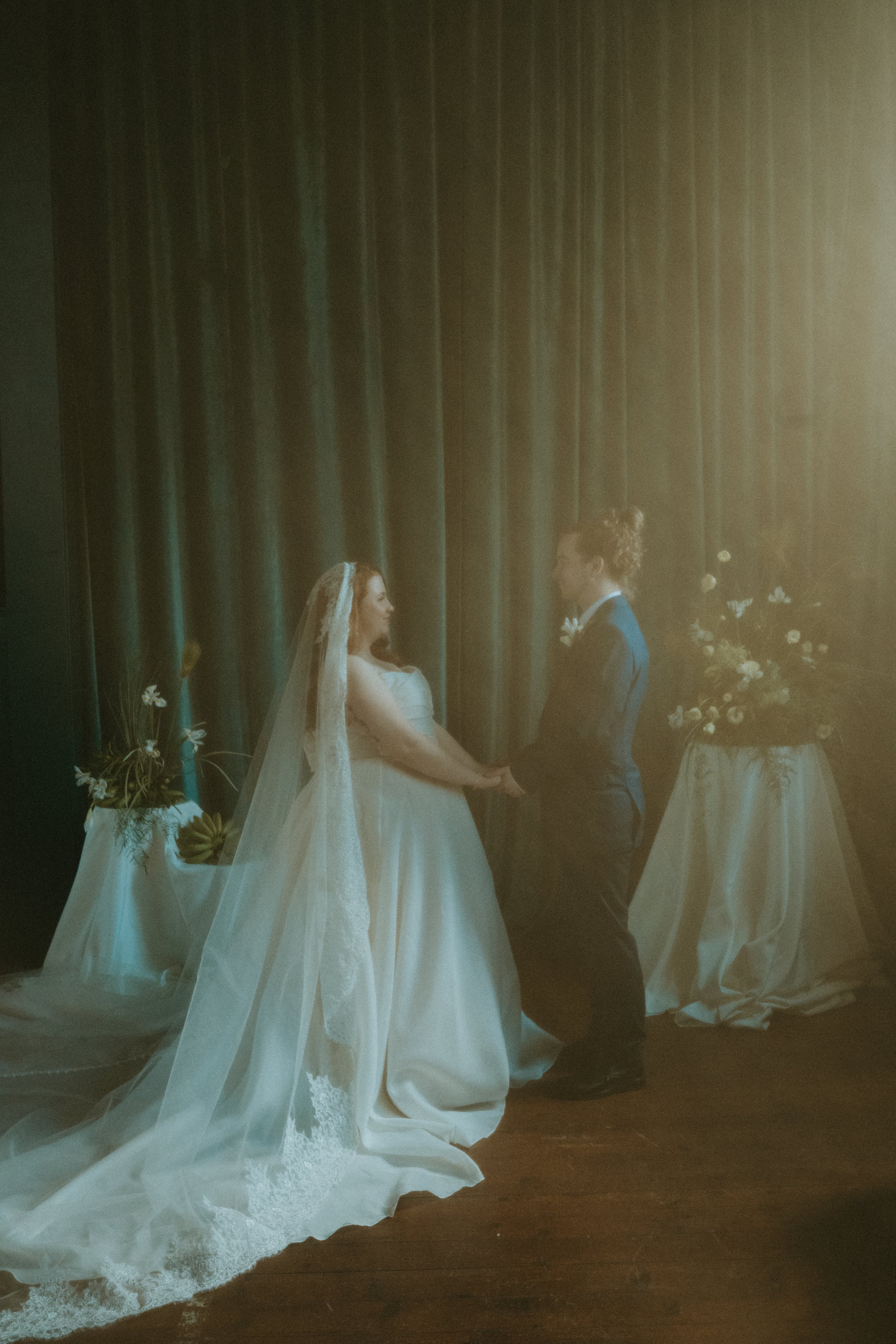A bride and groom holding hands during their wedding ceremony, standing in front of a dark curtain with floral arrangements on either side.