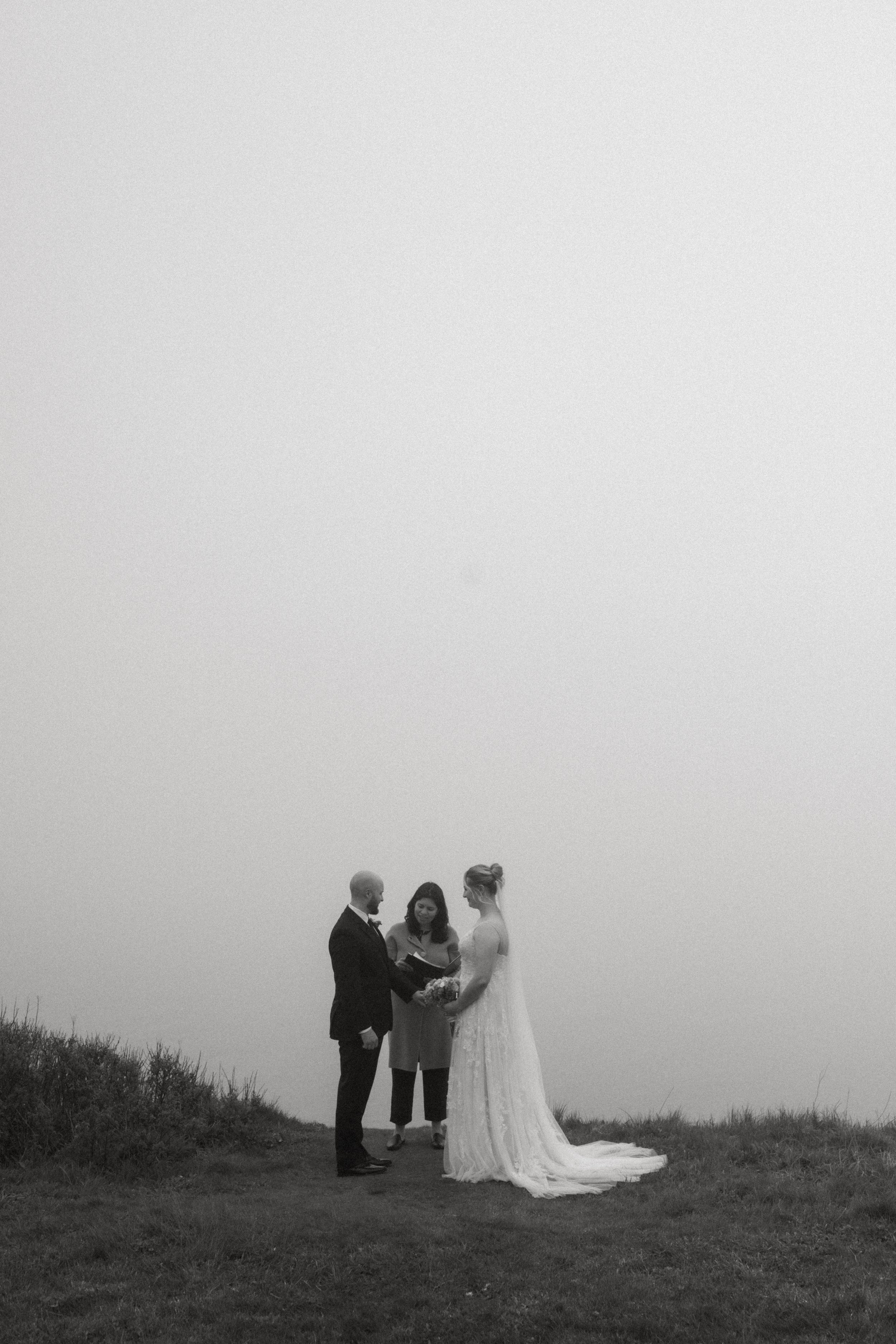 A wedding ceremony taking place outdoors on a grassy field with three people, a bride in a flowing white gown, a groom in a black suit, and an officiant in a coat, with a foggy sky background.