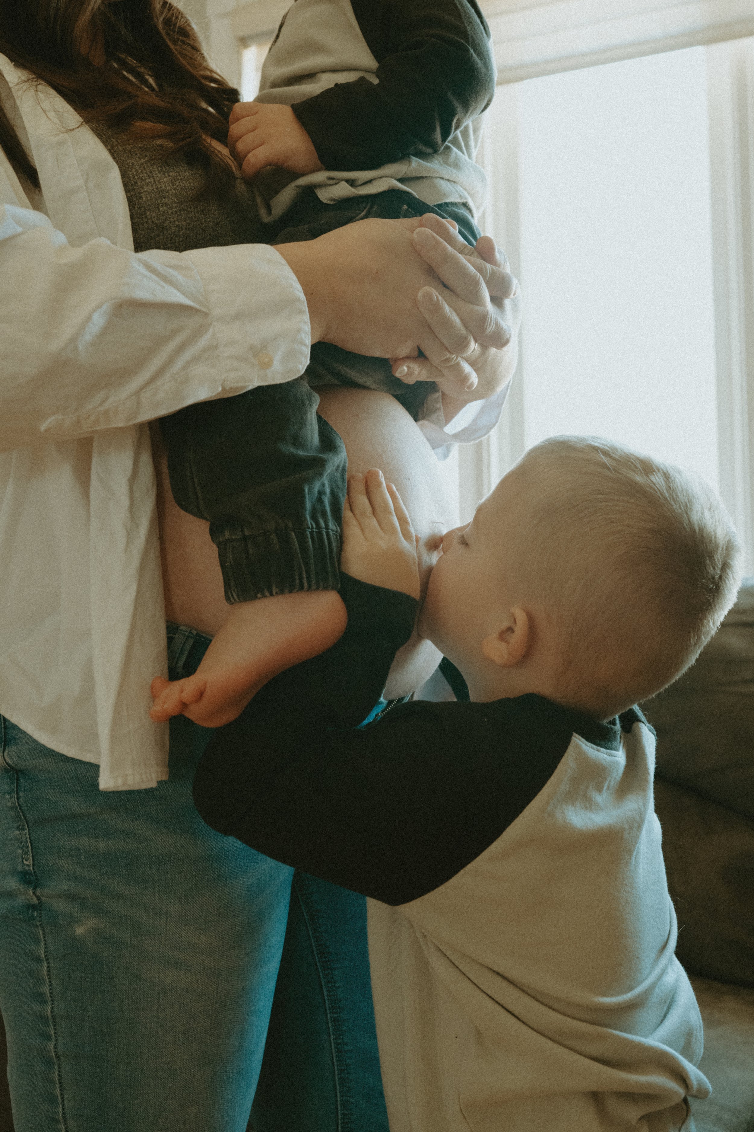 A young boy is kissing a pregnant woman's belly with a second child on her lap, while the woman is holding the younger child and standing near a window.