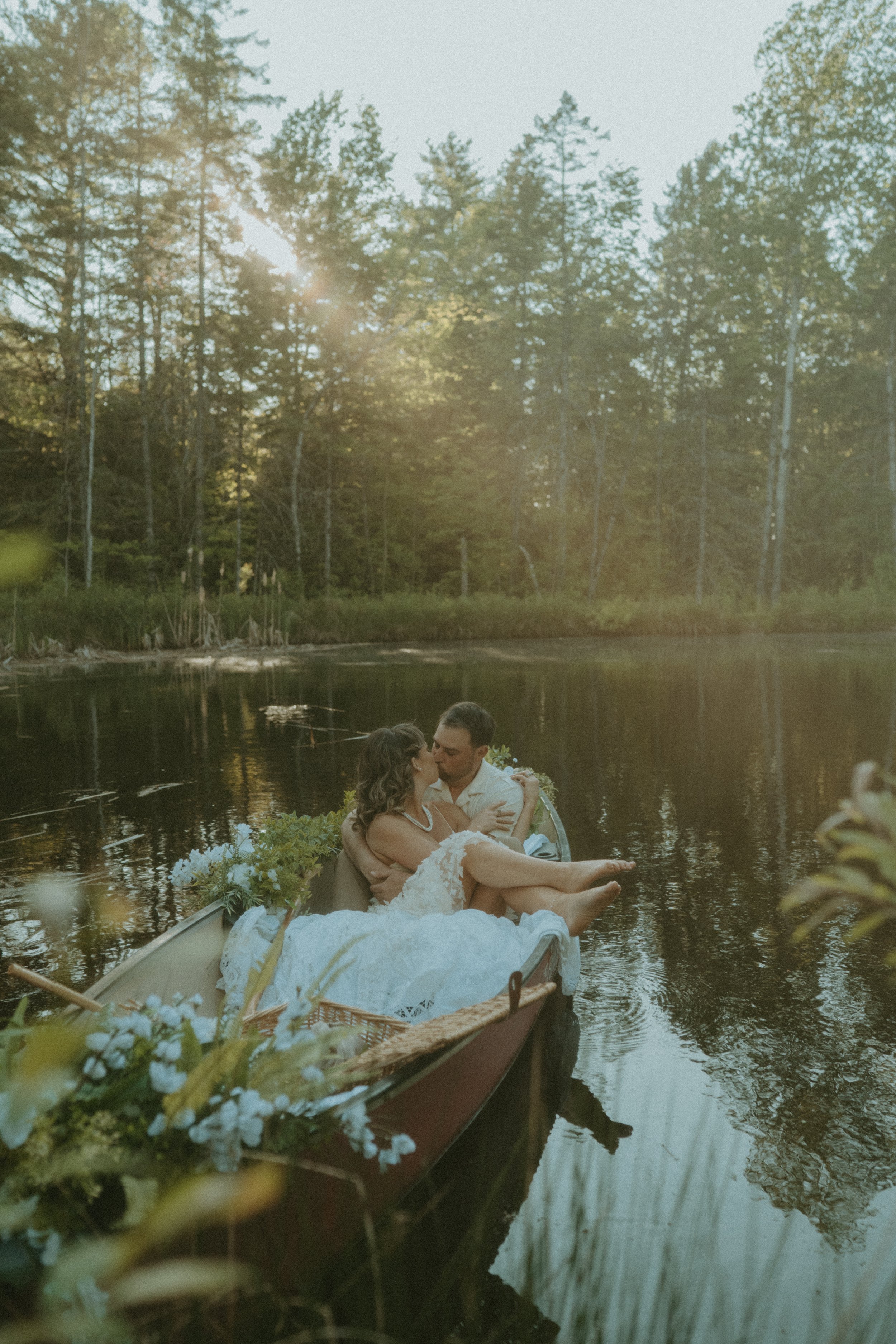 A couple in wedding attire sharing a kiss on a boat decorated with flowers, floating on a peaceful river surrounded by trees at sunset.