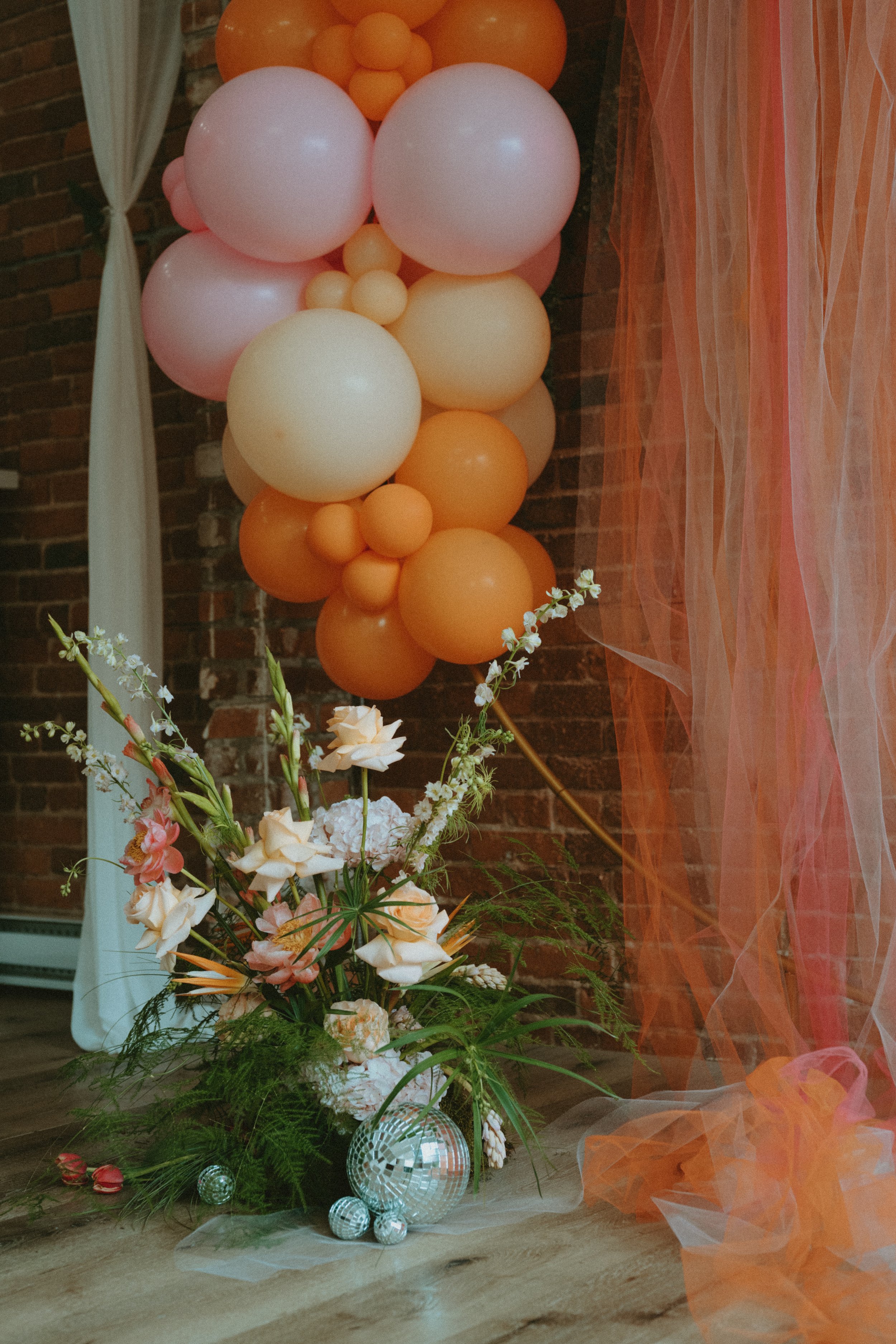 Decorative display featuring a large bouquet of pink and white flowers, surrounded by green foliage, with a cluster of pastel-colored balloons in soft pink, peach, and cream shades, set against a brick wall and sheer orange and pink fabric curtains.