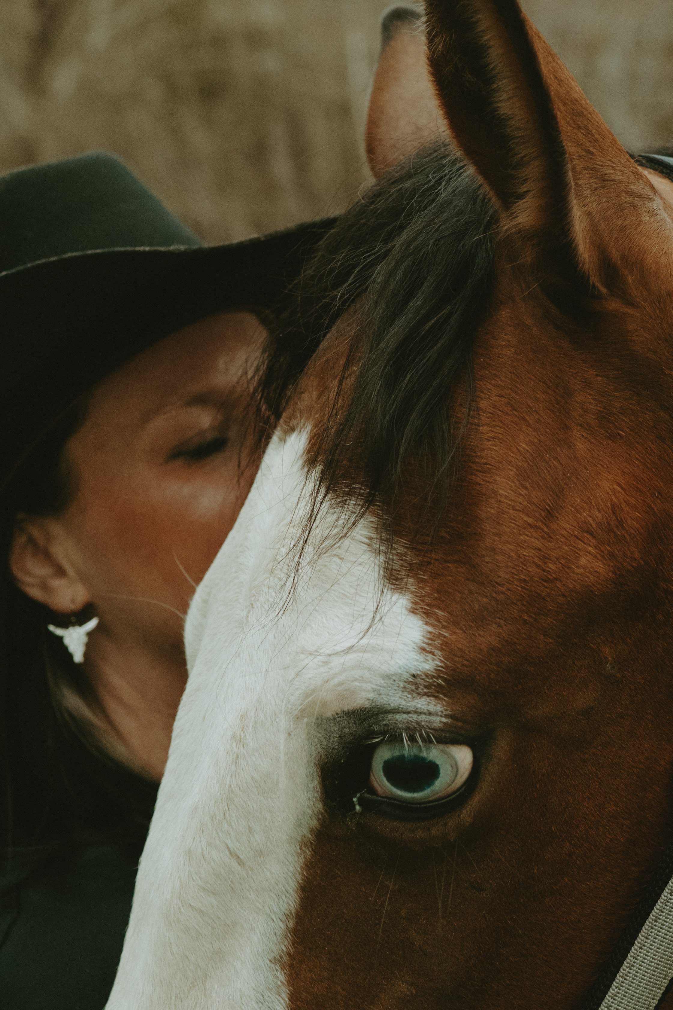 A woman with dark hair and earrings hugging a brown and white horse with blue eyes, close-up of their faces.