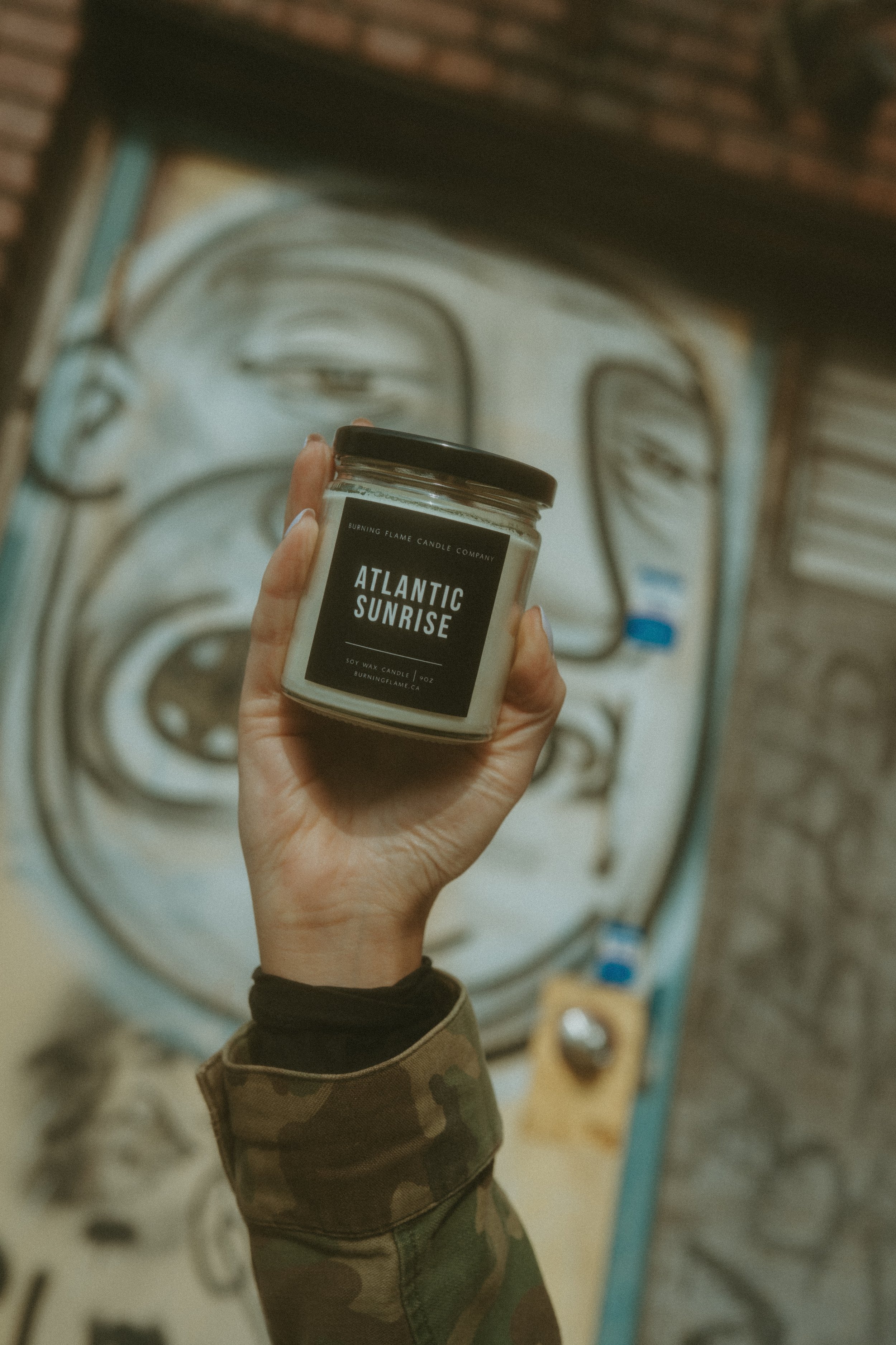 A hand holding a scented candle labeled Atlantic Sunrise in front of an abstract mural of a face.