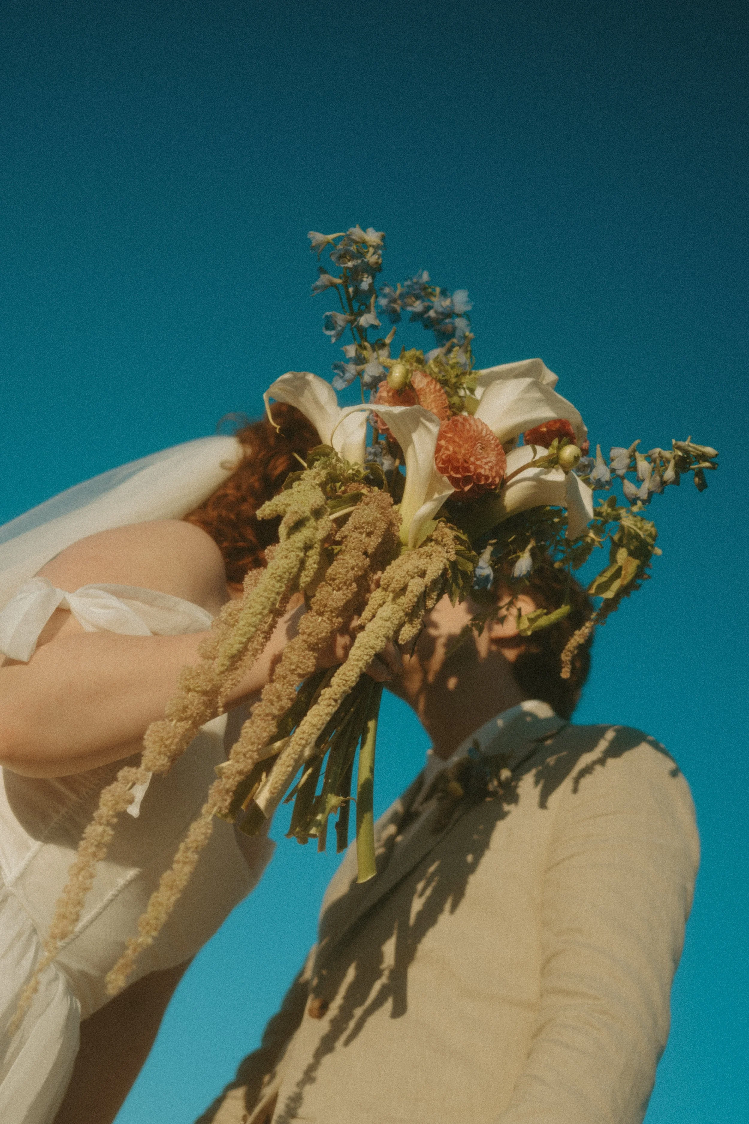 A person in a beige suit holding a bouquet of flowers in front of another person with a blue sky background taken by photos by devyn in Nova Scotia.