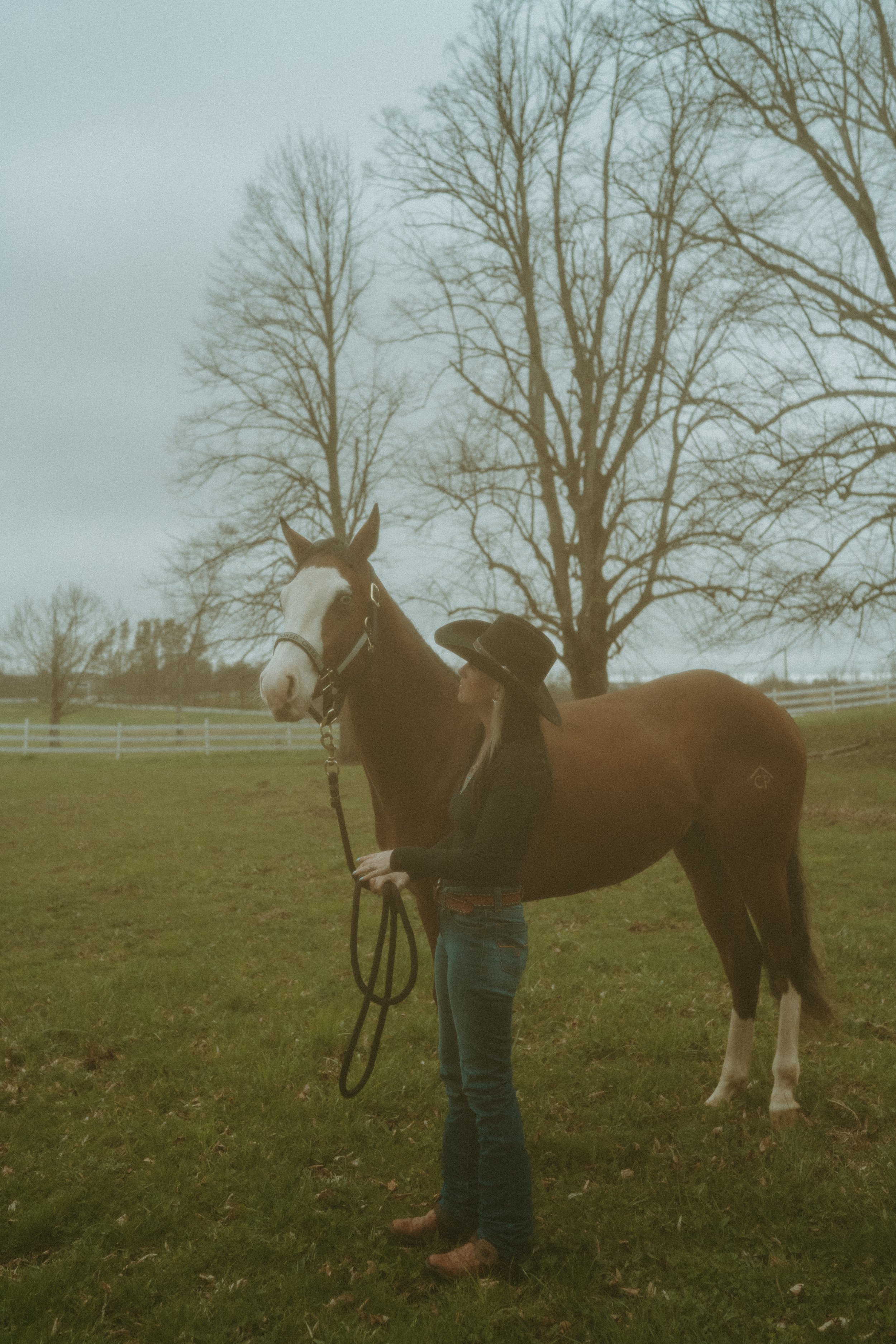 A person in a cowboy hat standing next to a brown and white horse on a grassy field with leafless trees in the background.