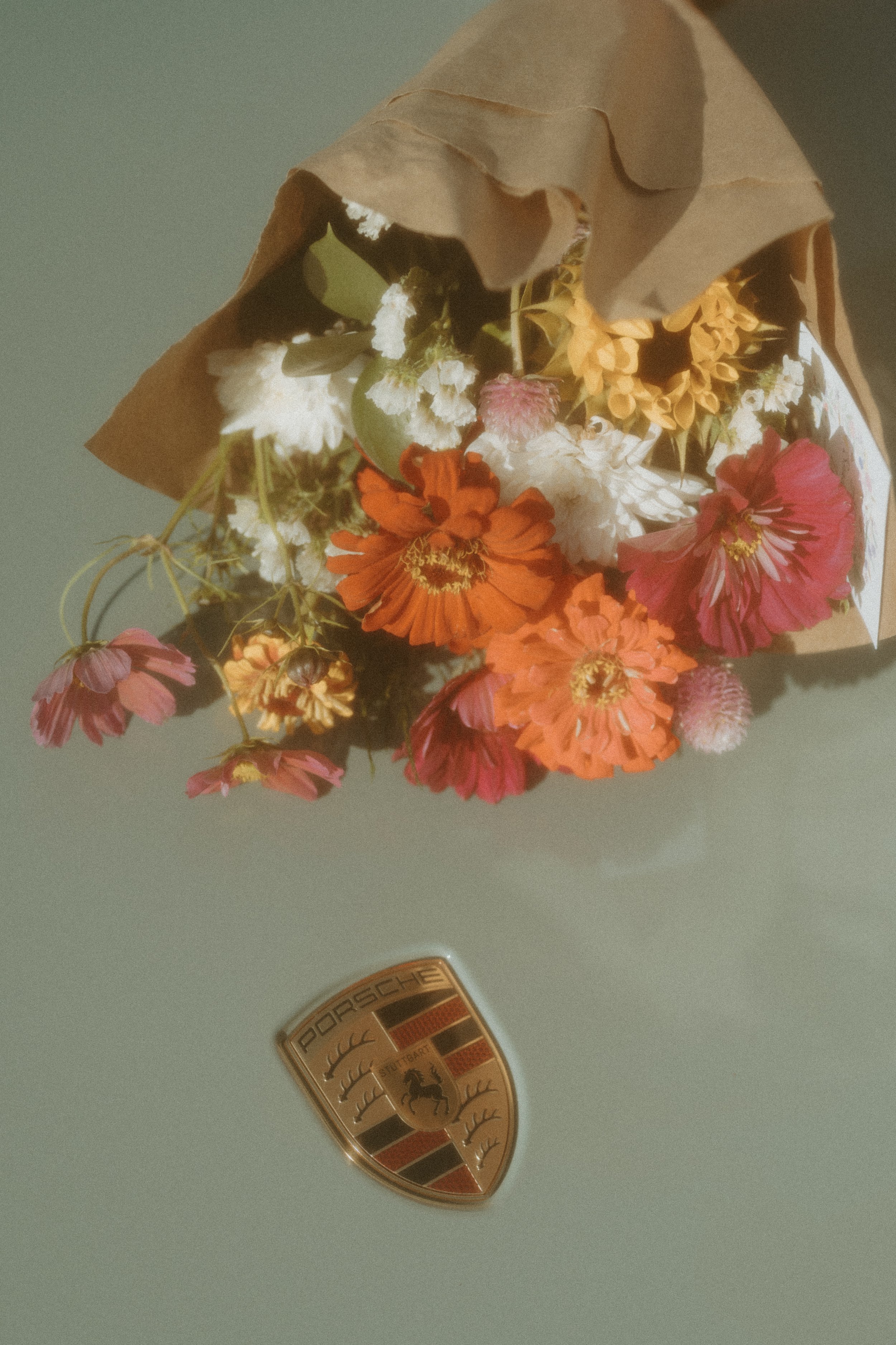 Bouquet of mixed flowers wrapped in brown paper laying on top of a light green Porsche car surface, showing Porsche logo.