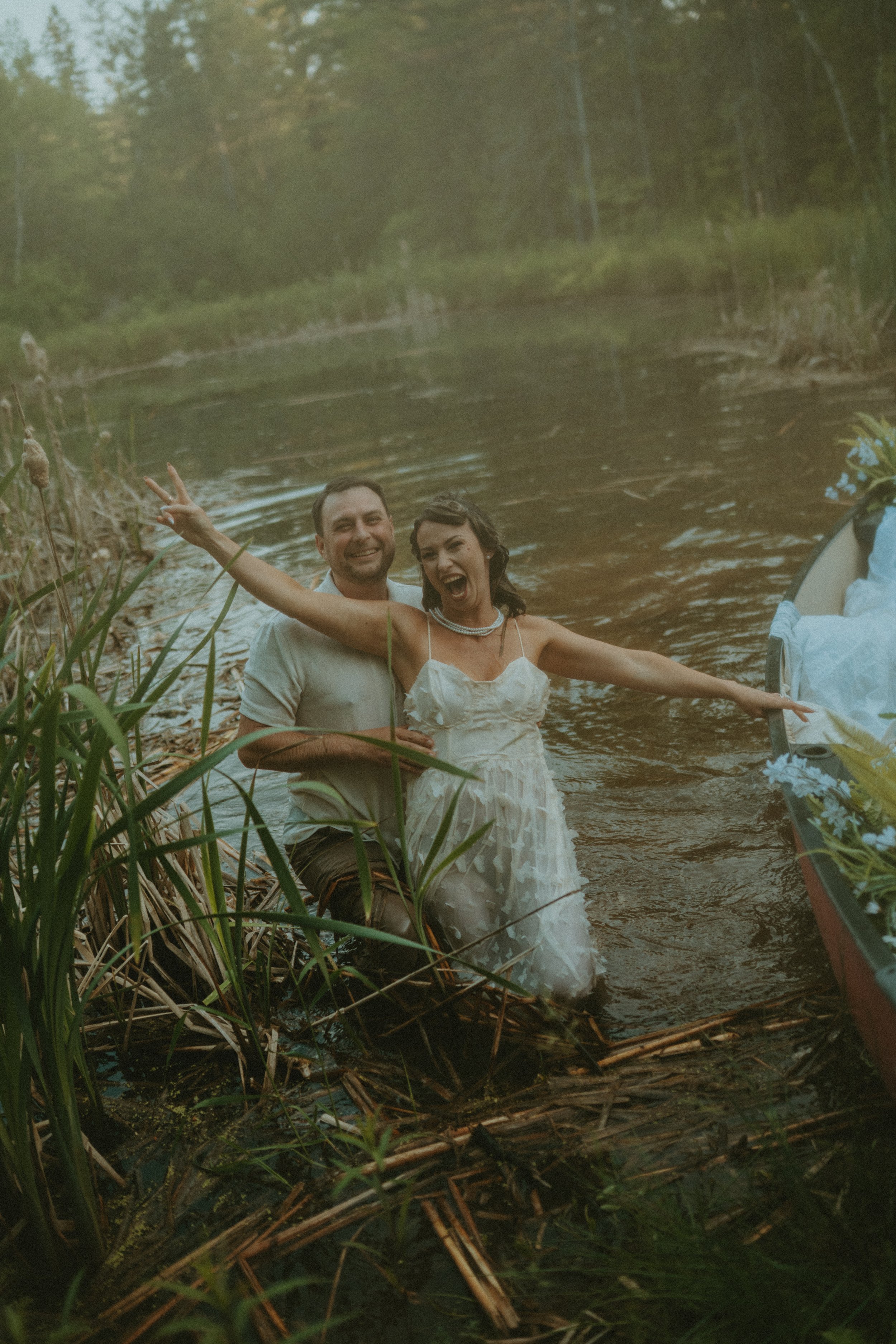 A couple dressed in wedding attire, standing in a shallow river, smiling and making peace signs, with a boat decorated with flowers nearby.