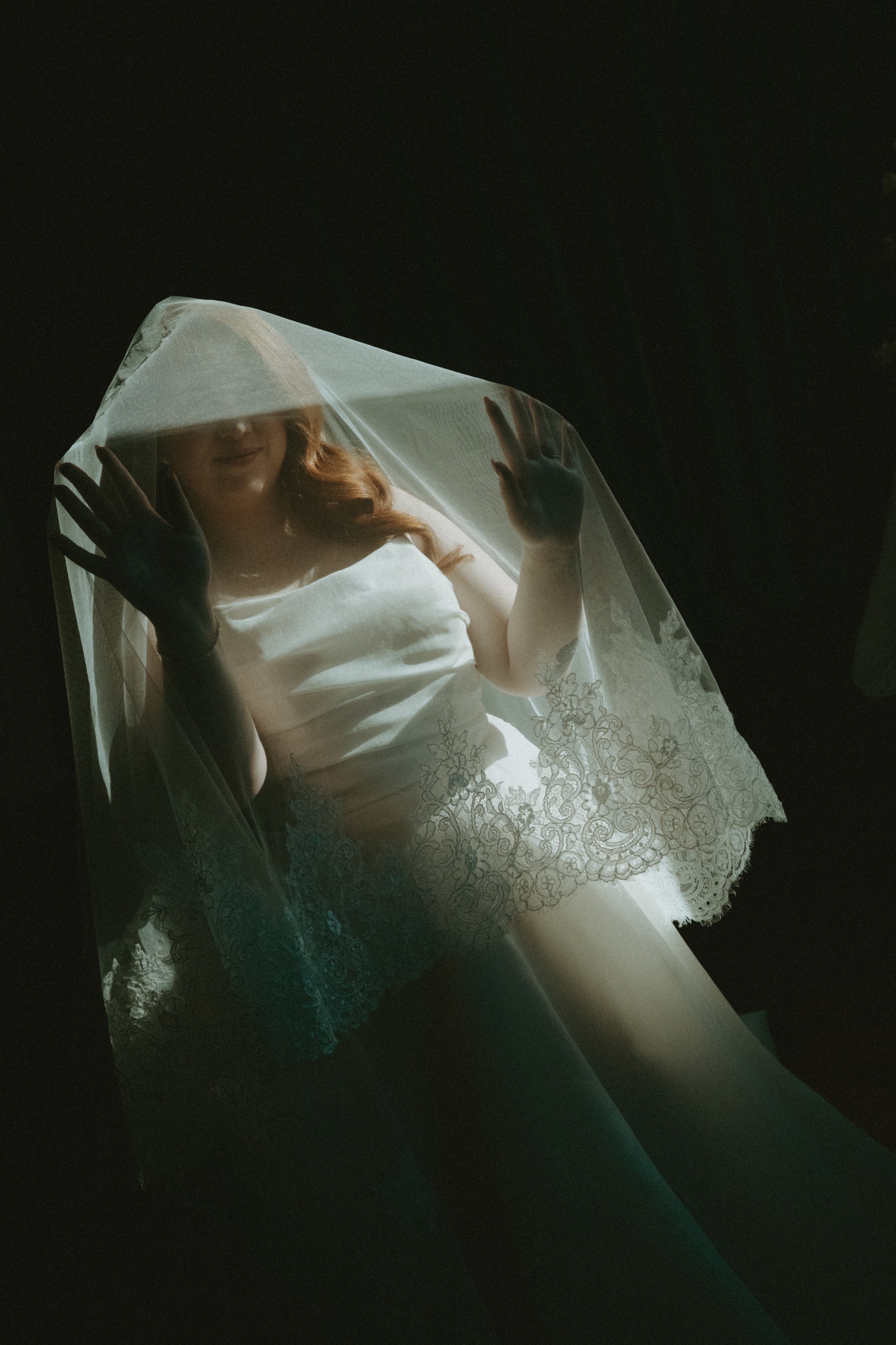 A woman in a lace-trimmed white dress, holding a sheer veil, looking up through it, with her face partially obscured in a dark setting.