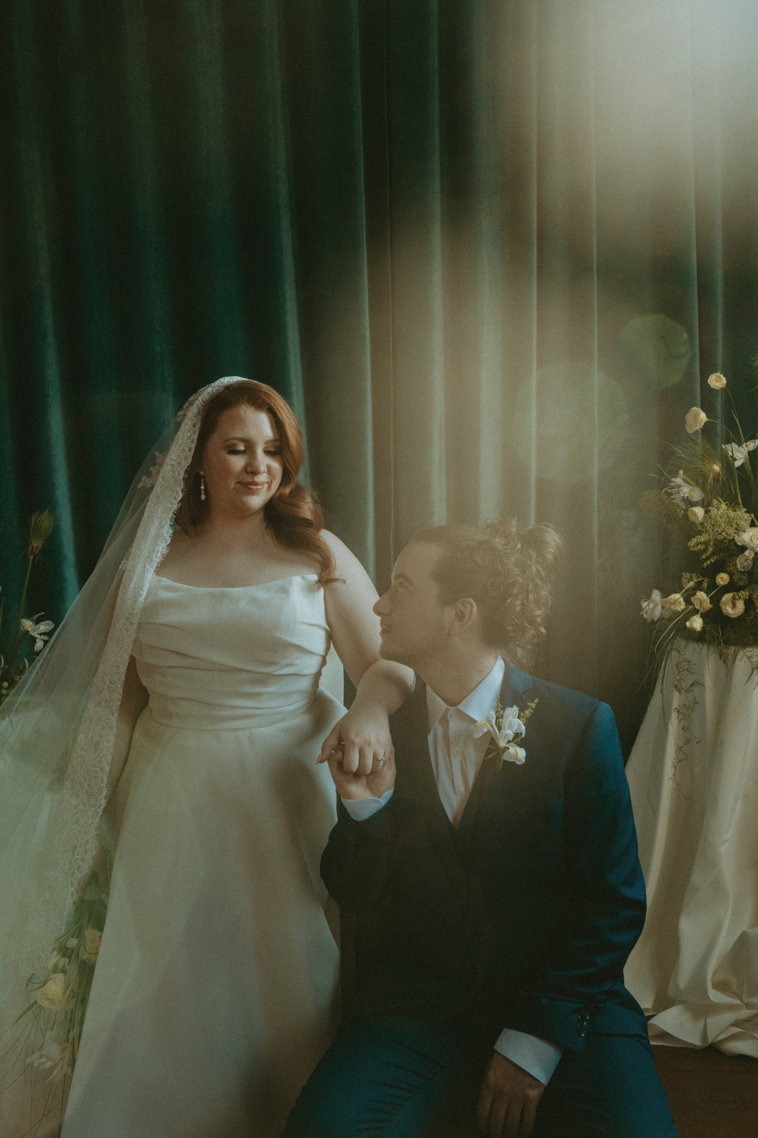 A bride and groom sharing a romantic moment indoors. The bride is wearing a white wedding dress and veil, and the groom is dressed in a dark suit with a white shirt. The bride is standing, and the groom is kneeling, holding her hand and looking up at
