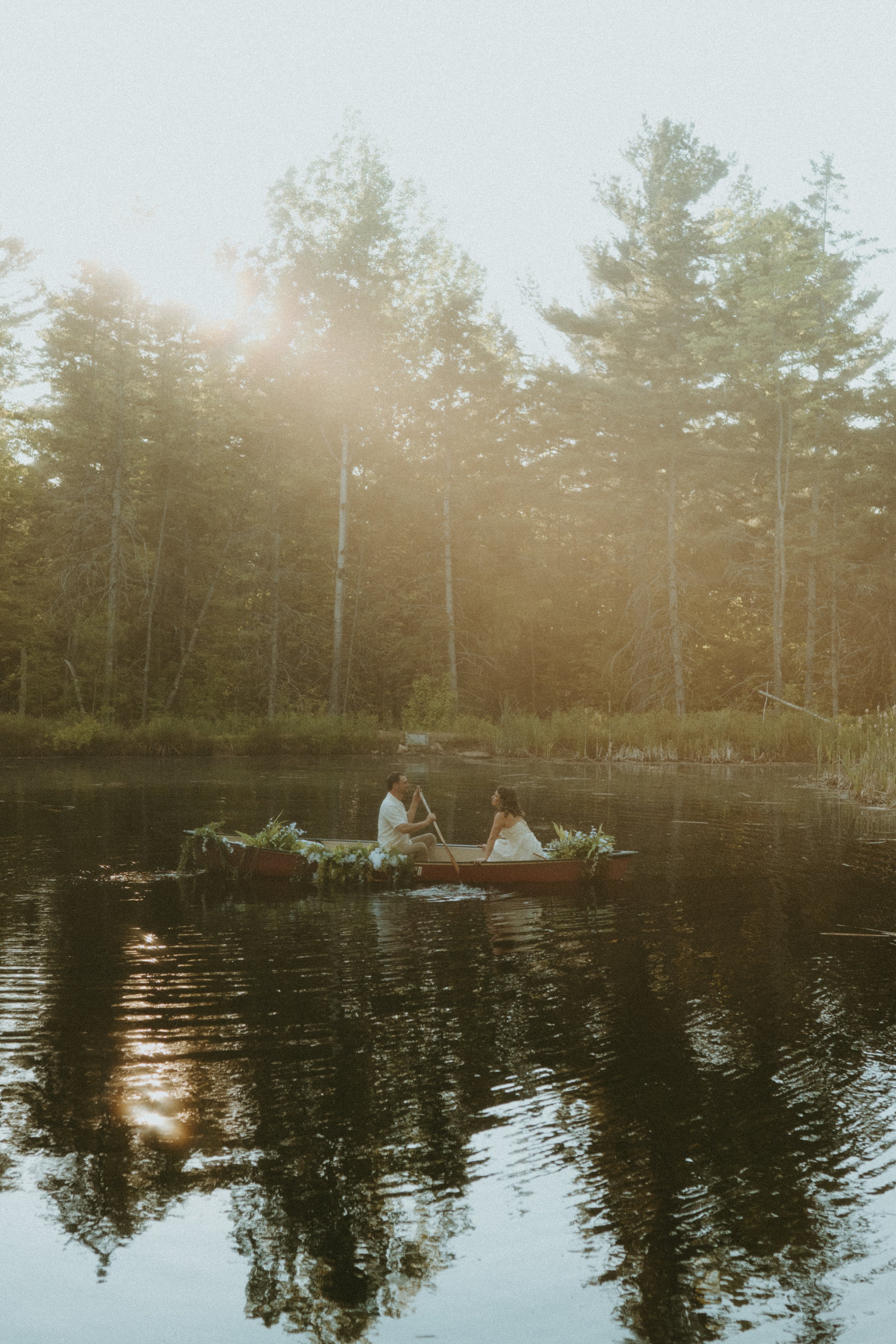 A couple in wedding attire on a boat decorated with flowers, floating on a calm lake surrounded by trees, with sunlight shining through the trees.