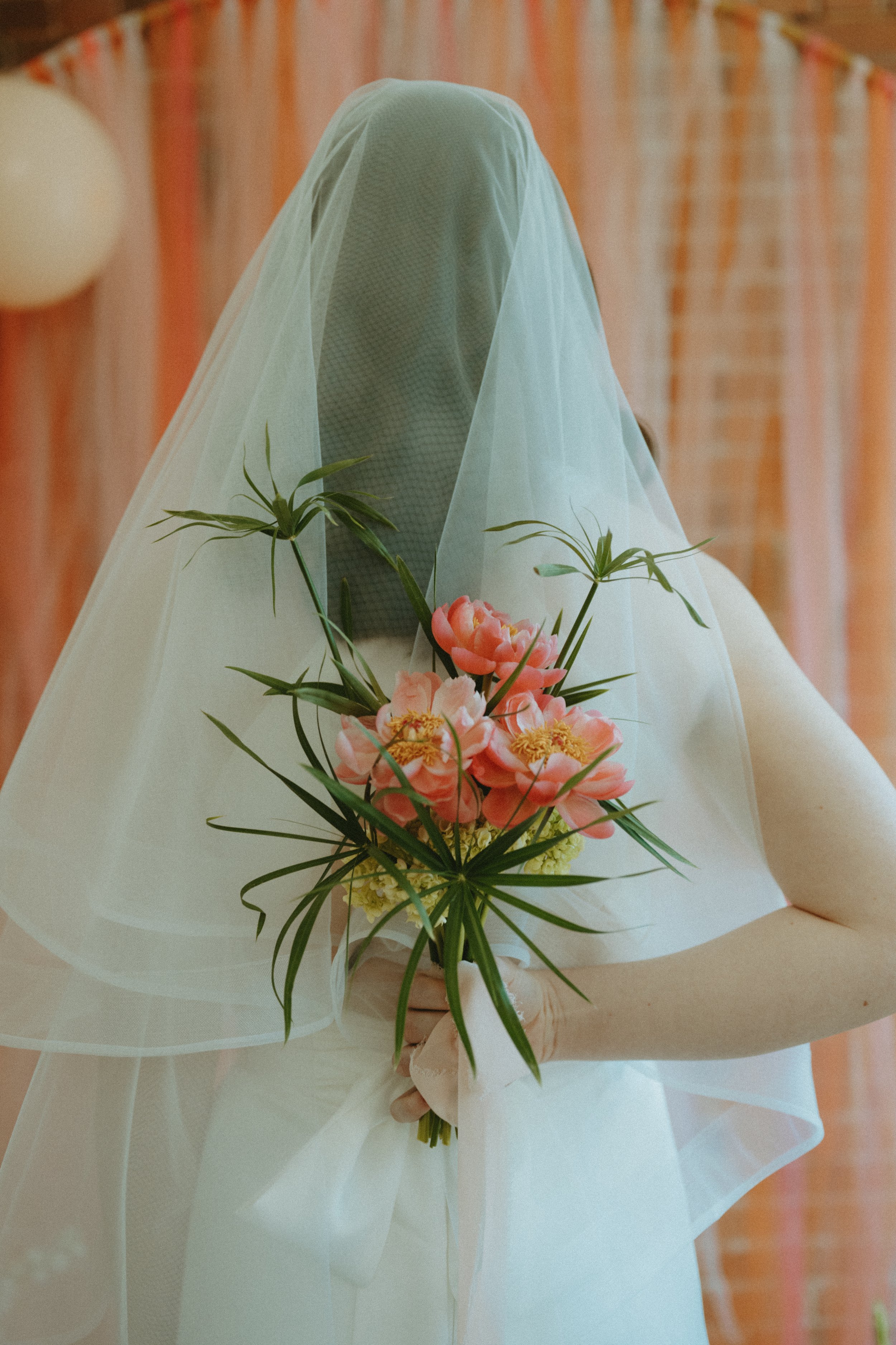 A person dressed in white holding a bouquet of pink flowers and greenery, with a sheer veil covering their face, in a decorated indoor setting.