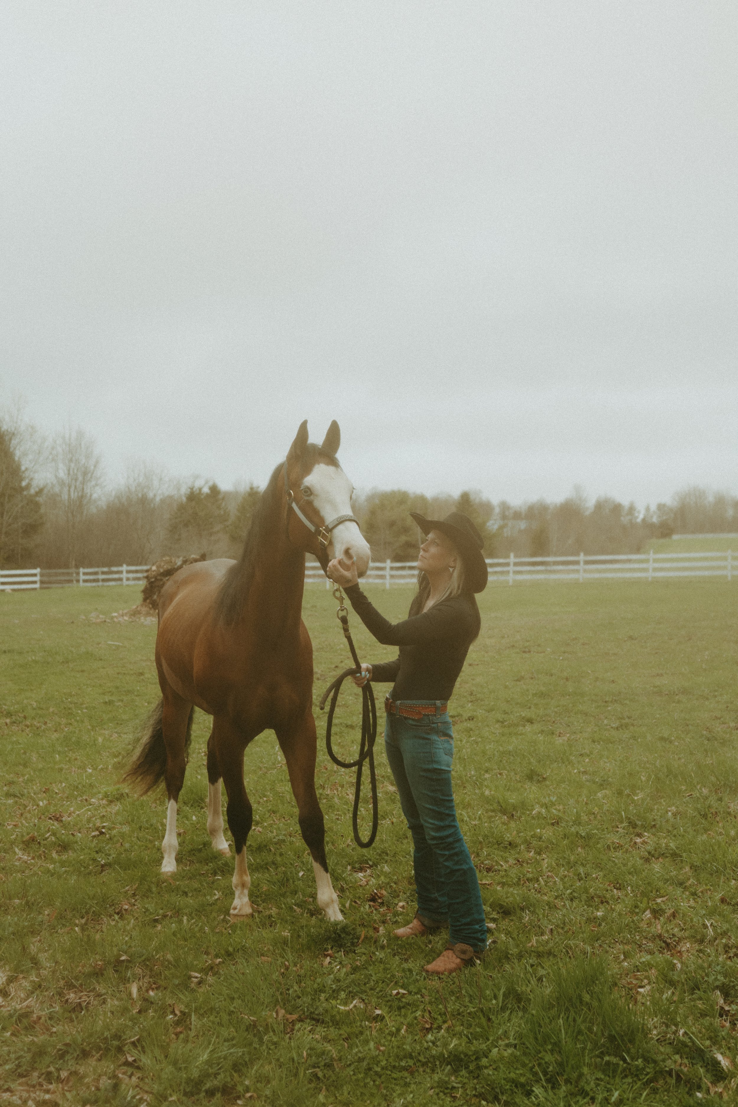 A woman in a cowboy hat and black shirt standing on green grass, holding the reins of a brown and white horse and gently touching its face.