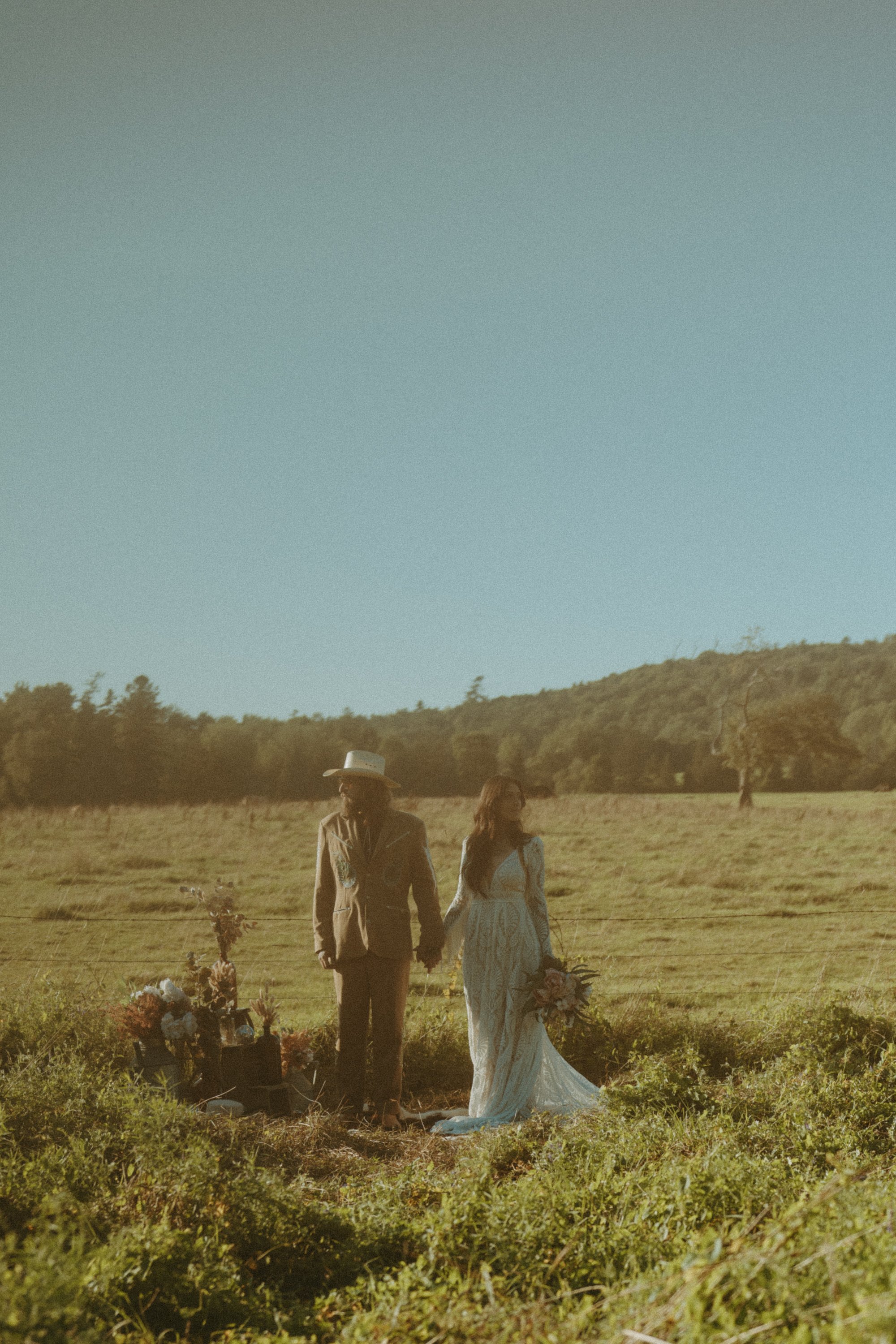 A couple standing outdoors in a grassy field with a clear blue sky, holding hands and dressed in wedding attire. The woman wears a long white dress and holds a bouquet, while the man wears a cowboy hat, a suit, and glasses. Flowers are arranged on th