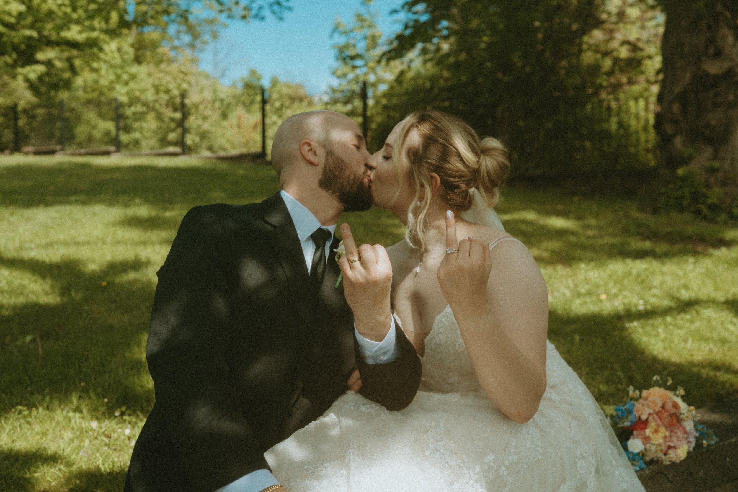 A newlywed couple sharing a kiss outside on a sunny day. The bride is wearing a white wedding dress with lace details and has her hair styled in an updo. The groom is in a black suit and tie. They are sitting on the grass with a colorful bouquet of f