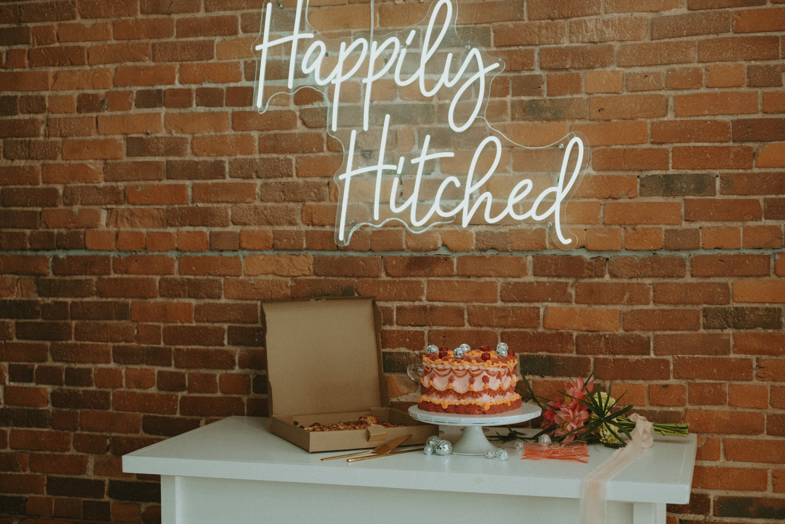 Decorative setup with a neon sign reading 'Happily Hitched' on a brick wall, a cake on a stand, a pizza box, flowers, and some scattered decorations on a white table.