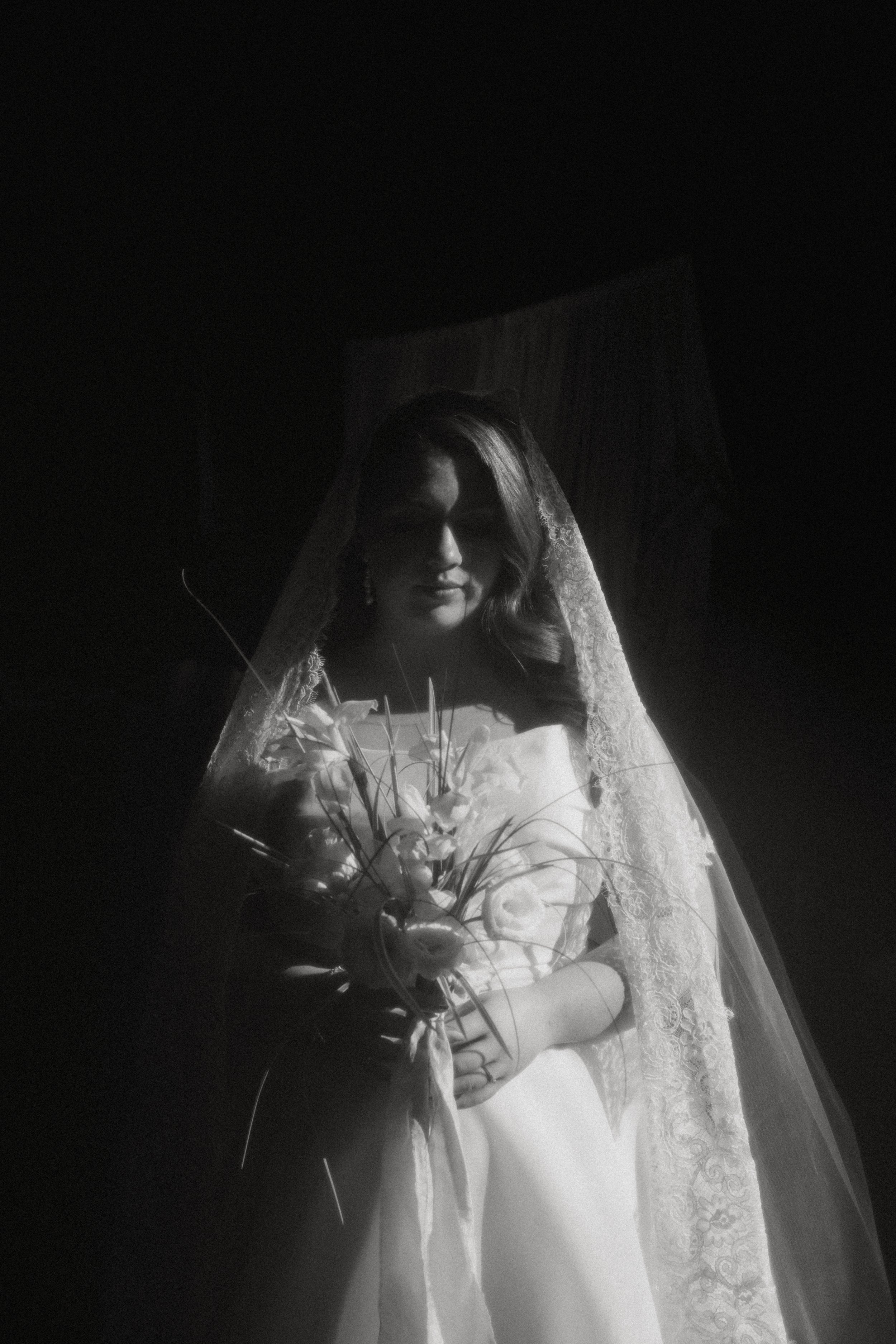 Black and white photo of a bride holding a bouquet, wearing a veil and wedding dress, standing in a dimly lit room.