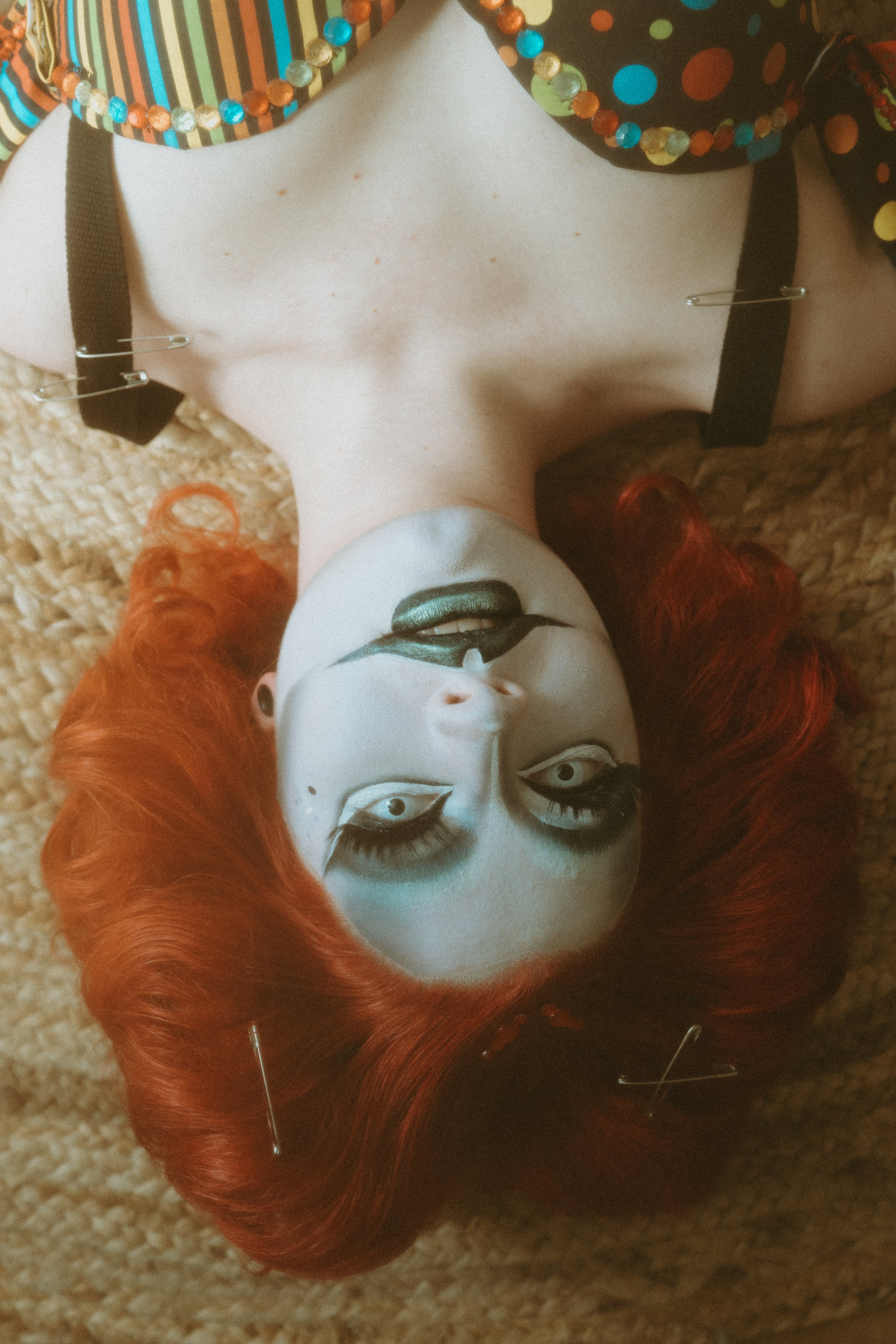 A person with red hair lying on a textured surface, with dramatic makeup including black eyeliner, blue eyeshadow, and metallic green lips, wearing a colorful polka dot bra with safety pins and a striped top with beads.