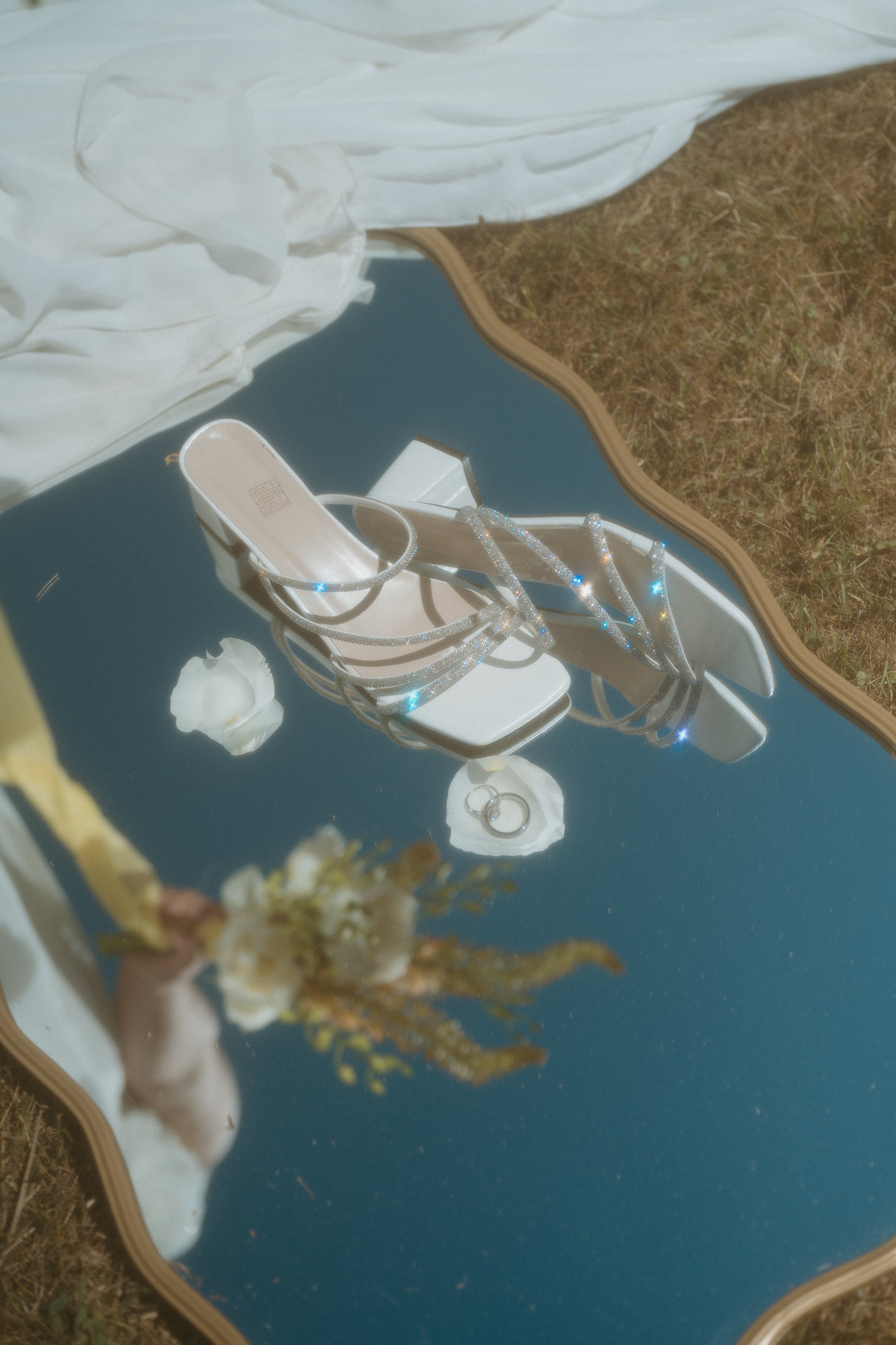 A pair of white, strappy high-heeled sandals with sparkly embellishments, reflected in a mirror placed on the grass. A small bouquet of flowers is held by a person taking the photo, with the sky and grass also visible.
