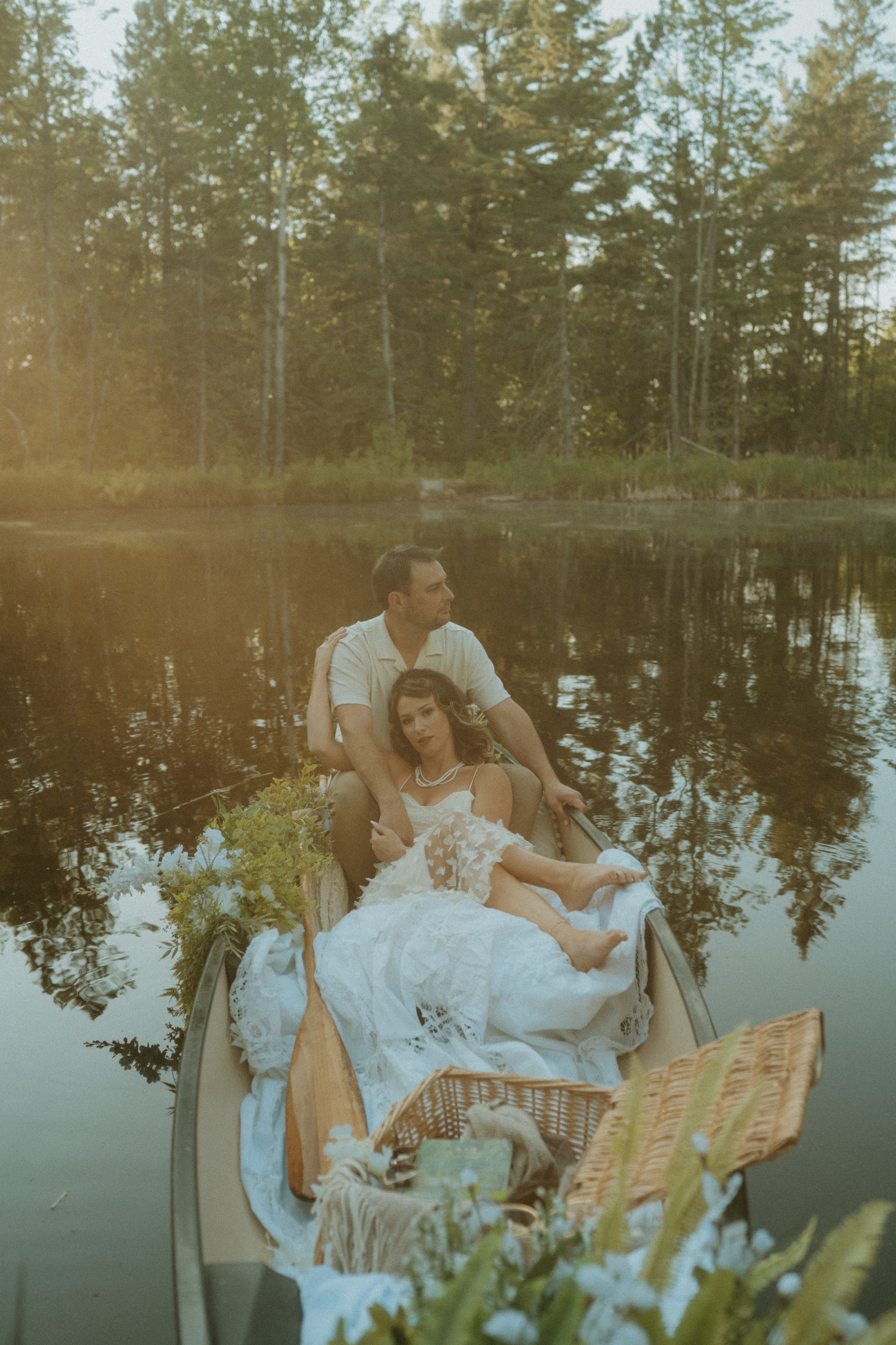 A man and a woman in wedding attire sitting in a boat on a calm river surrounded by trees, with golden sunlight filtering through the foliage.