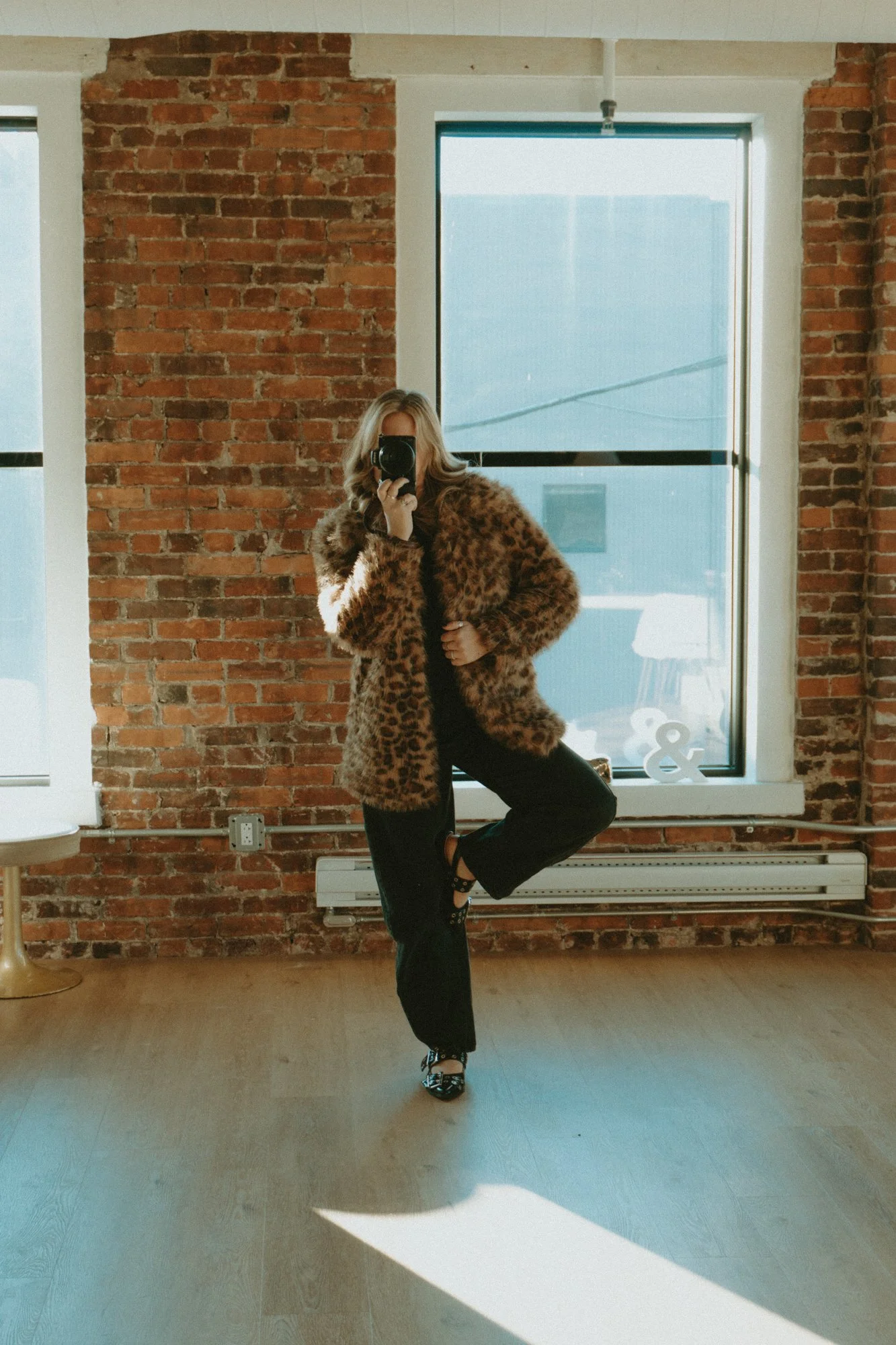 A woman wearing a leopard print fur coat, black pants with side buttons, and leopard print heels, taking a mirror selfie in a room with exposed brick walls and large windows. Sunlight is streaming into the room. Photo taken by Photos By Devyn.