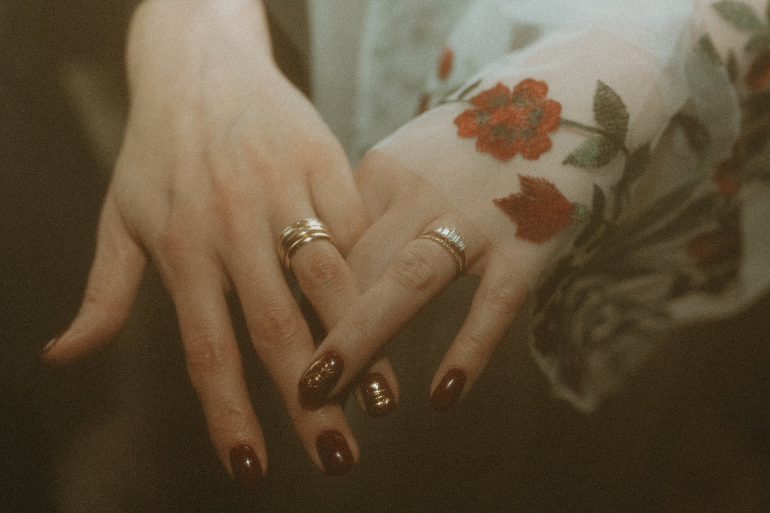Close-up of two hands with rings, one hand resting on top of the other, on a dark surface. One hand has dark red nail polish, and the other has gold rings. The person is wearing a sheer blouse with floral embroidery featuring red and orange flowers.