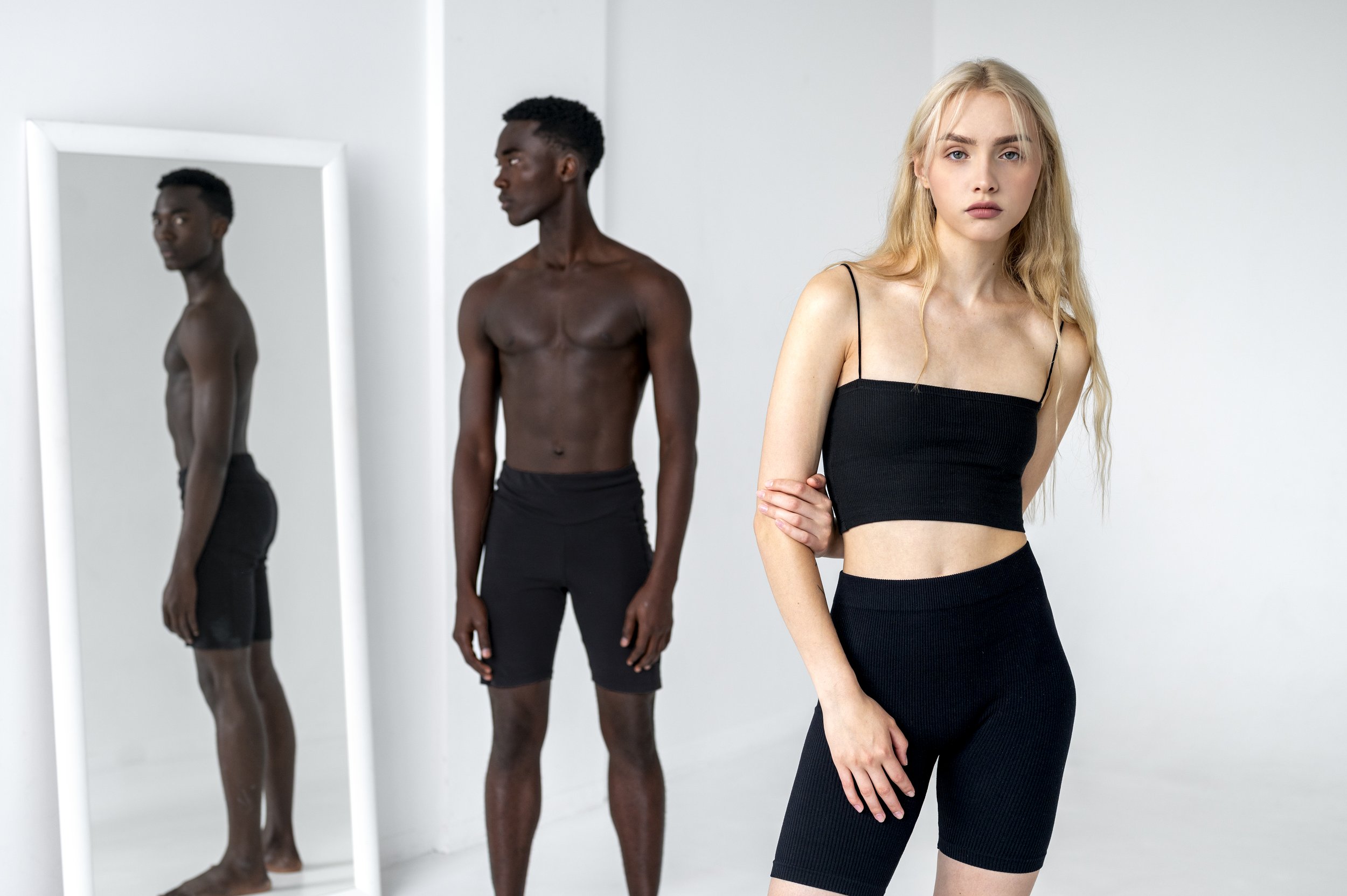 A blonde woman in a black crop top and high-waisted shorts standing in front of a mirror. Two shirtless men with dark skin and black shorts are reflected in the mirror, one looking away and the other looking directly at the camera.