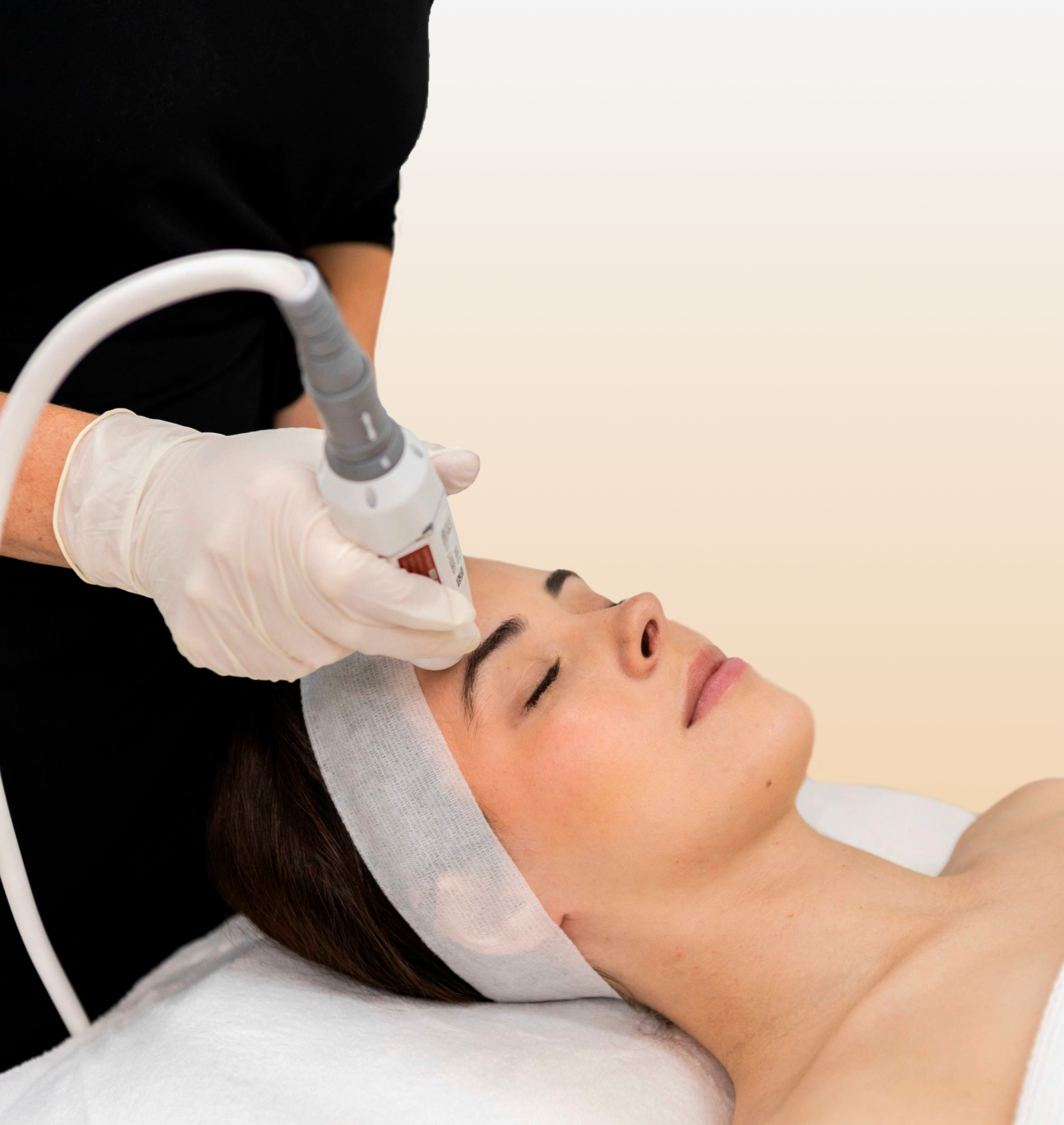 A woman receives a facial treatment with a modern skincare device in a spa or clinic setting, lying on a bed with a headband, while a professional in gloves operates the device.