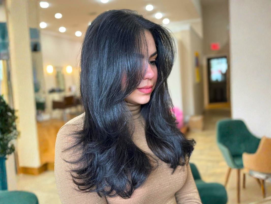 A woman with long black hair and a tan turtleneck sweater sitting in a cafe or restaurant with green chairs and warm lighting.