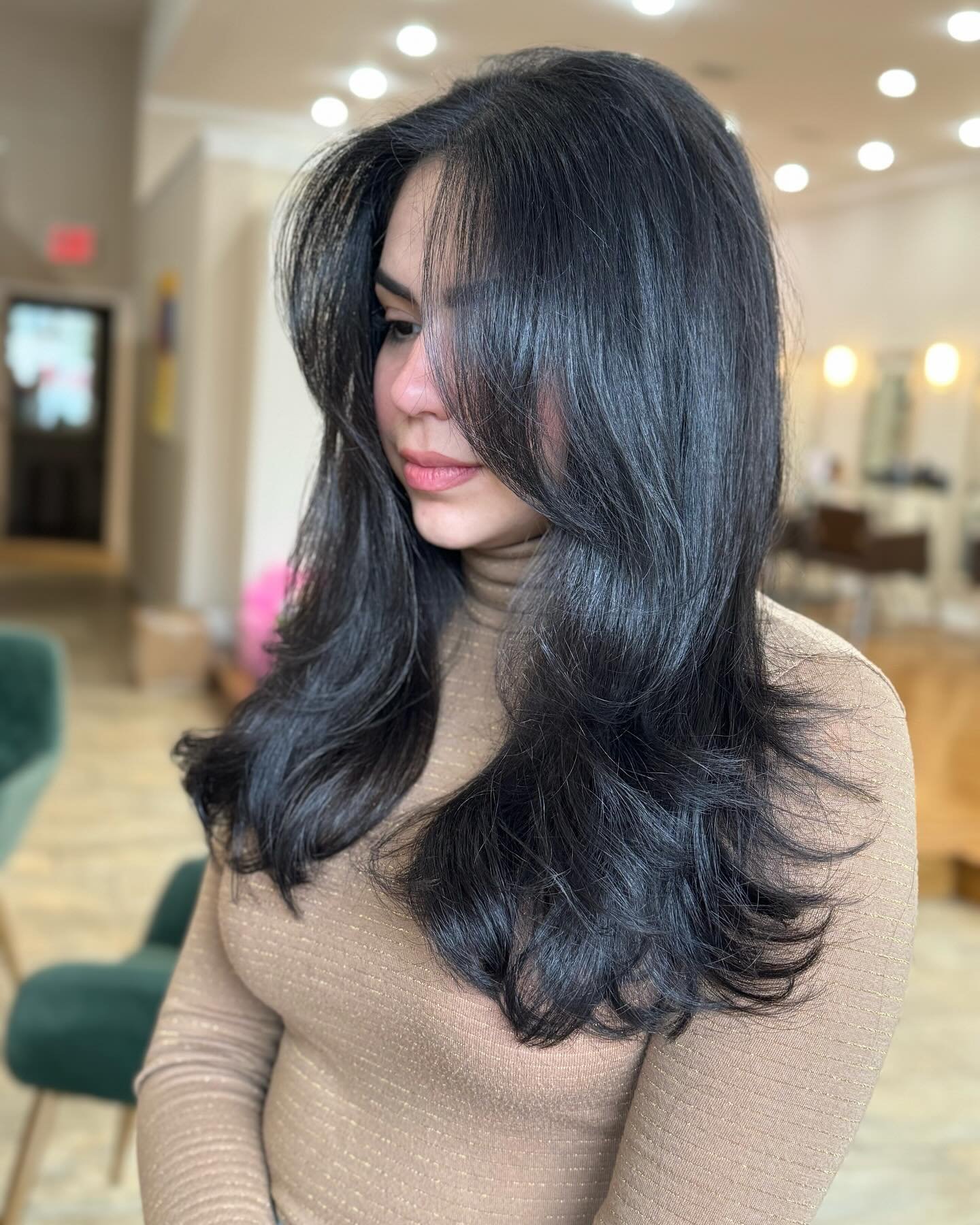 Woman with long, shiny black hair styled in loose waves, wearing a beige, textured long-sleeve top, standing indoors with blurred warm lighting and furniture in the background.