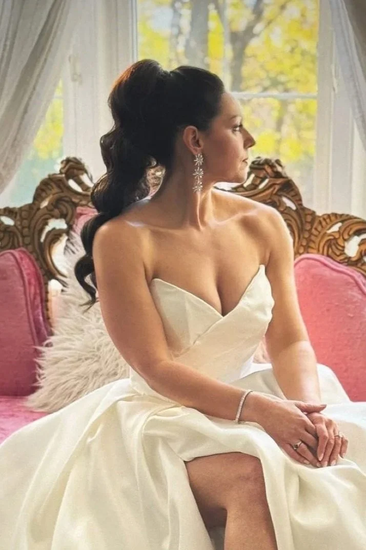 A woman in a strapless wedding dress sitting on a vintage pink sofa with carved wooden details, her hair in an elegant updo, wearing chandelier earrings, sitting by a window with sheer curtains, and looking away.