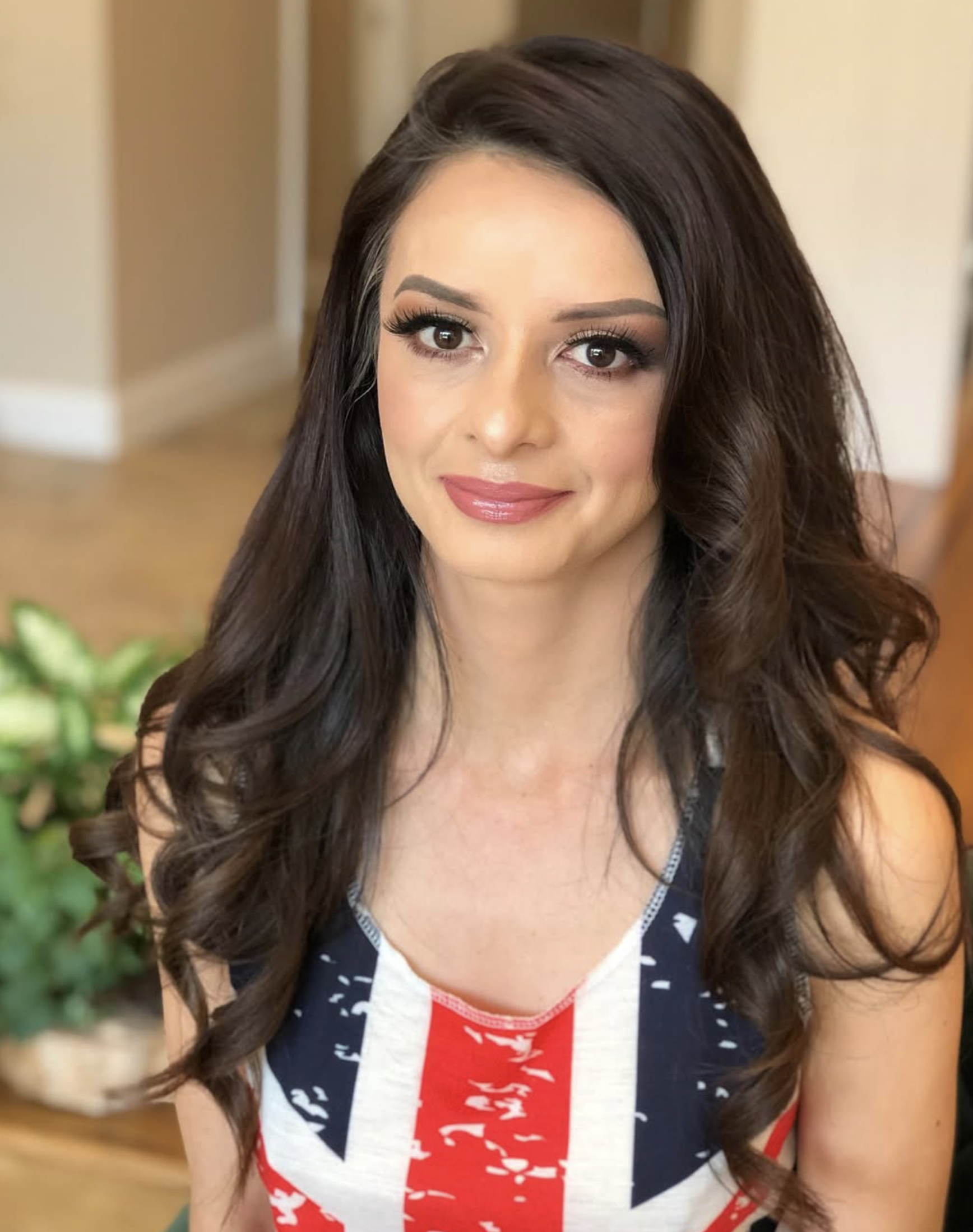 A woman with long, dark, wavy hair and makeup, wearing a sports jersey with red, white, and blue colors, smiling in a well-lit indoor setting.