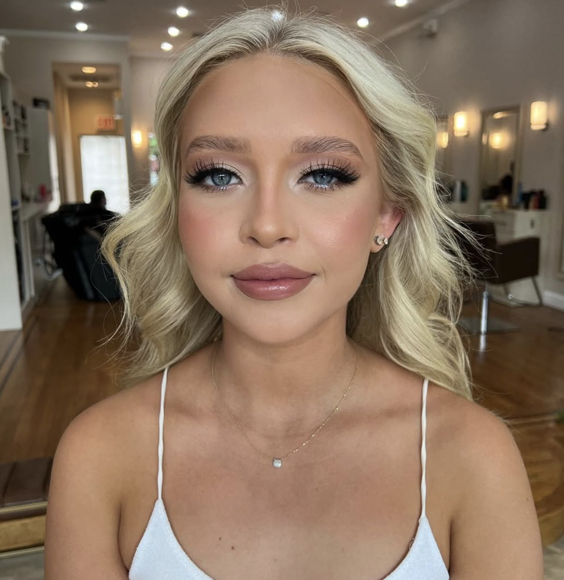 Close-up of a woman with blonde hair, blue eyes, and earrings, wearing a white top and a delicate necklace, in an indoor setting with warm lighting.