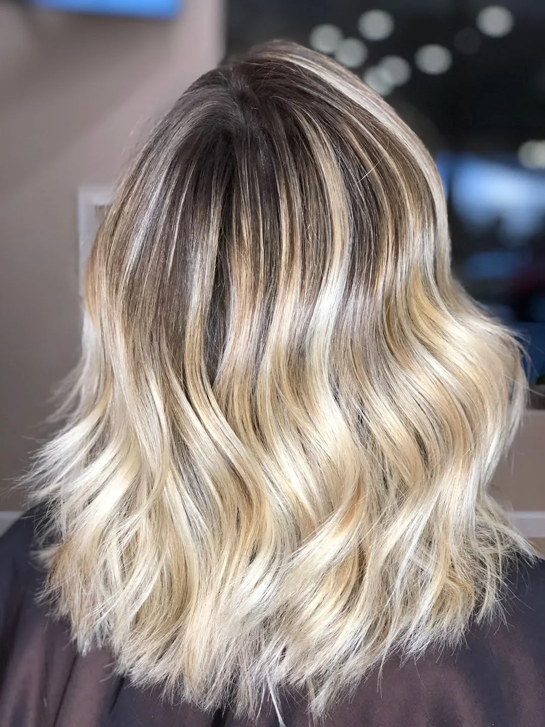 Back view of a woman's shoulder-length blonde wavy hair, a result of blonde balayage treatment.