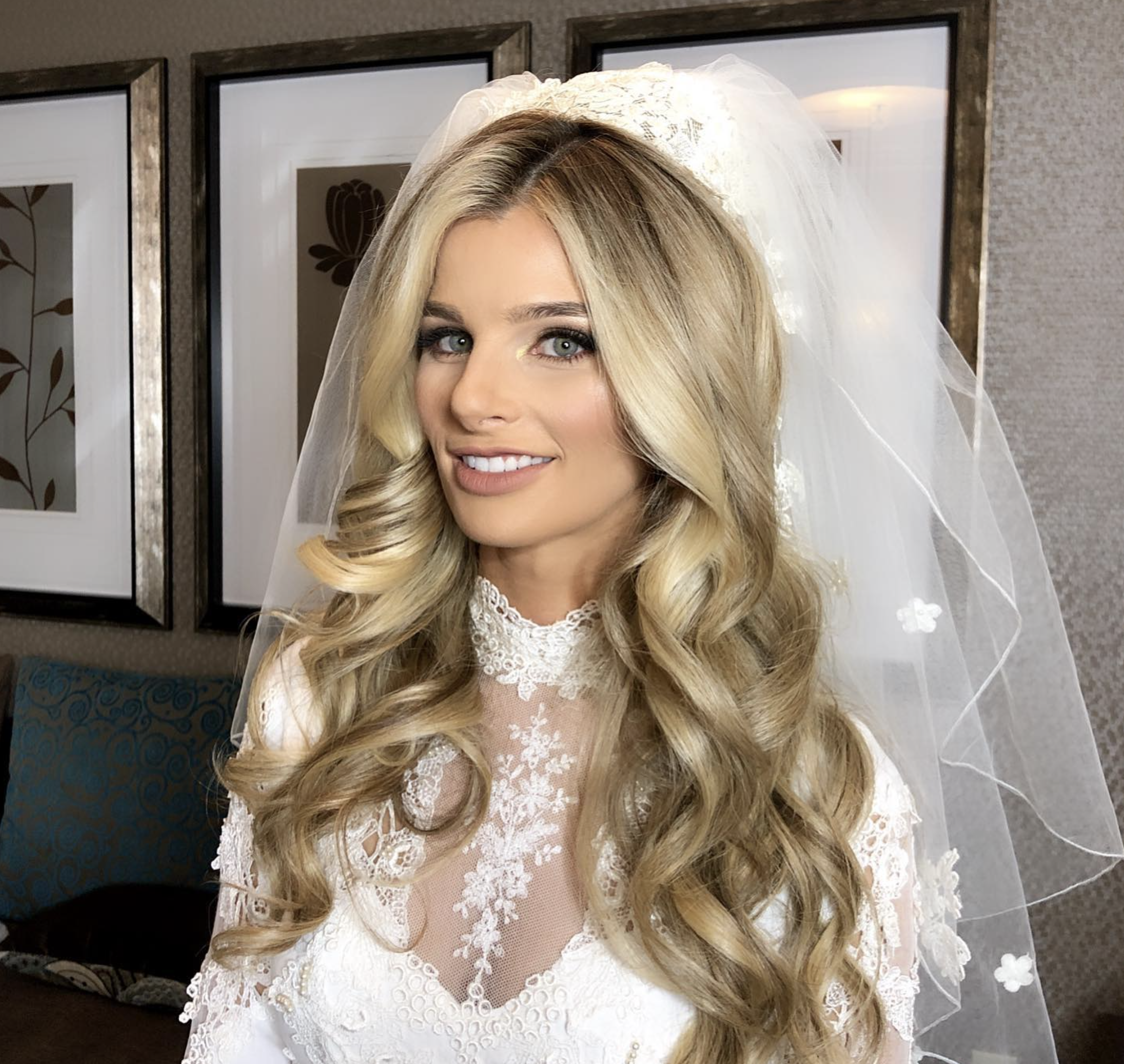 Close-up of a woman with blonde hair and blue eyes wearing a white lace wedding dress and veil, indoors with framed artwork on the wall in the background.